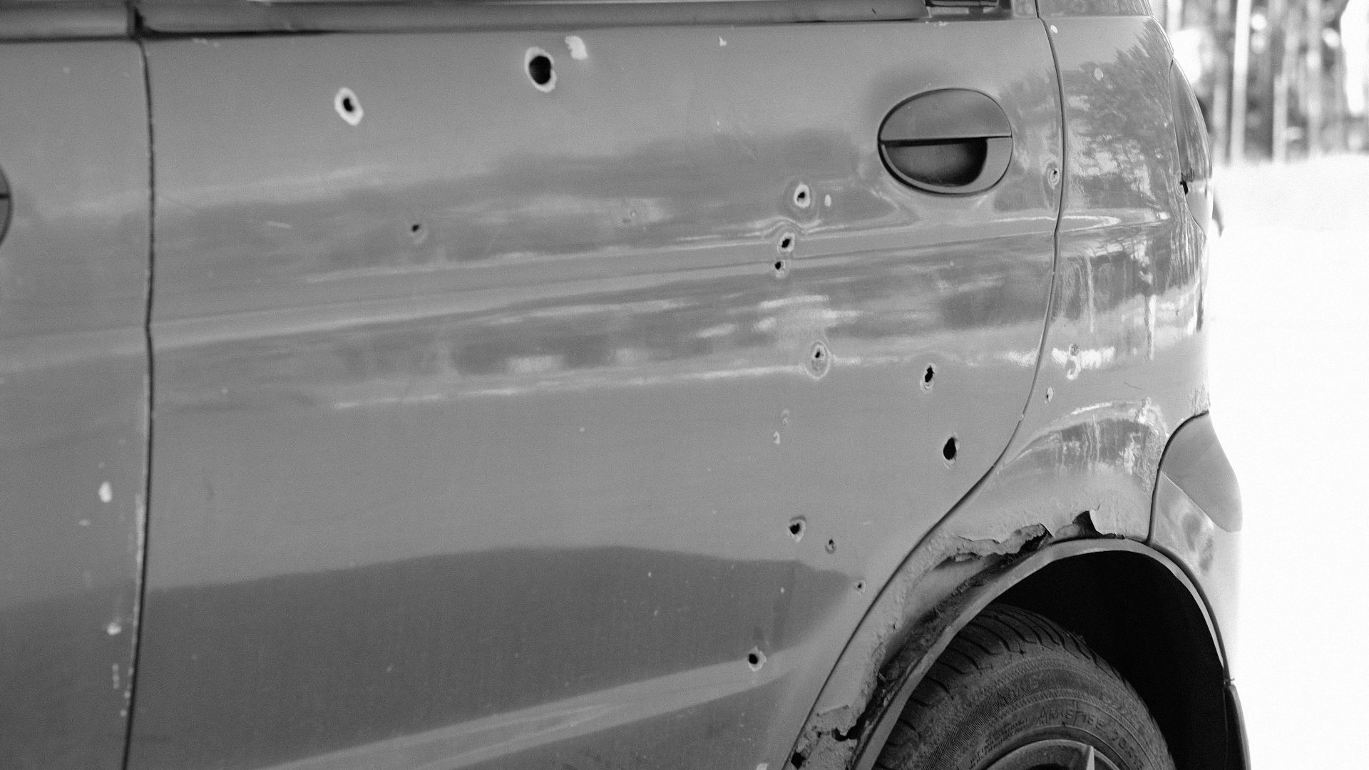 Close-up of a car door with bullet holes and a flat tire in a forest setting.