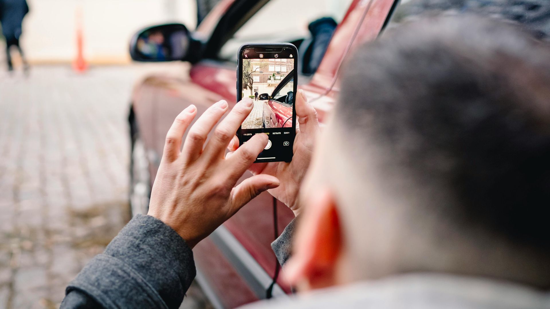 Man using smartphone to take pictures of an urban scene beside a red car.