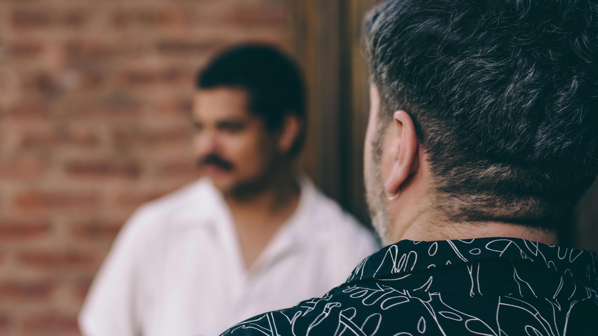 Two men engaged in conversation with a blurred focus against a brick wall background.