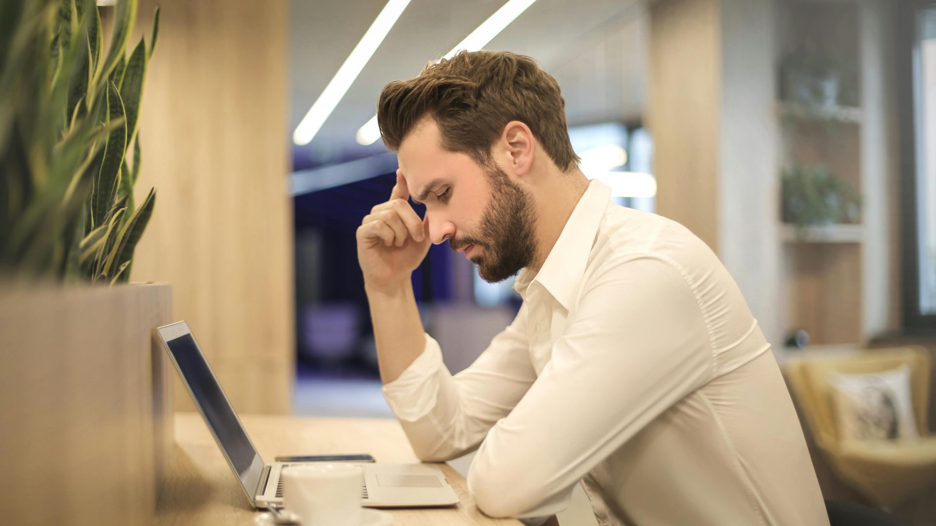 A thoughtful man in a shirt works on his laptop at a modern indoor office space.