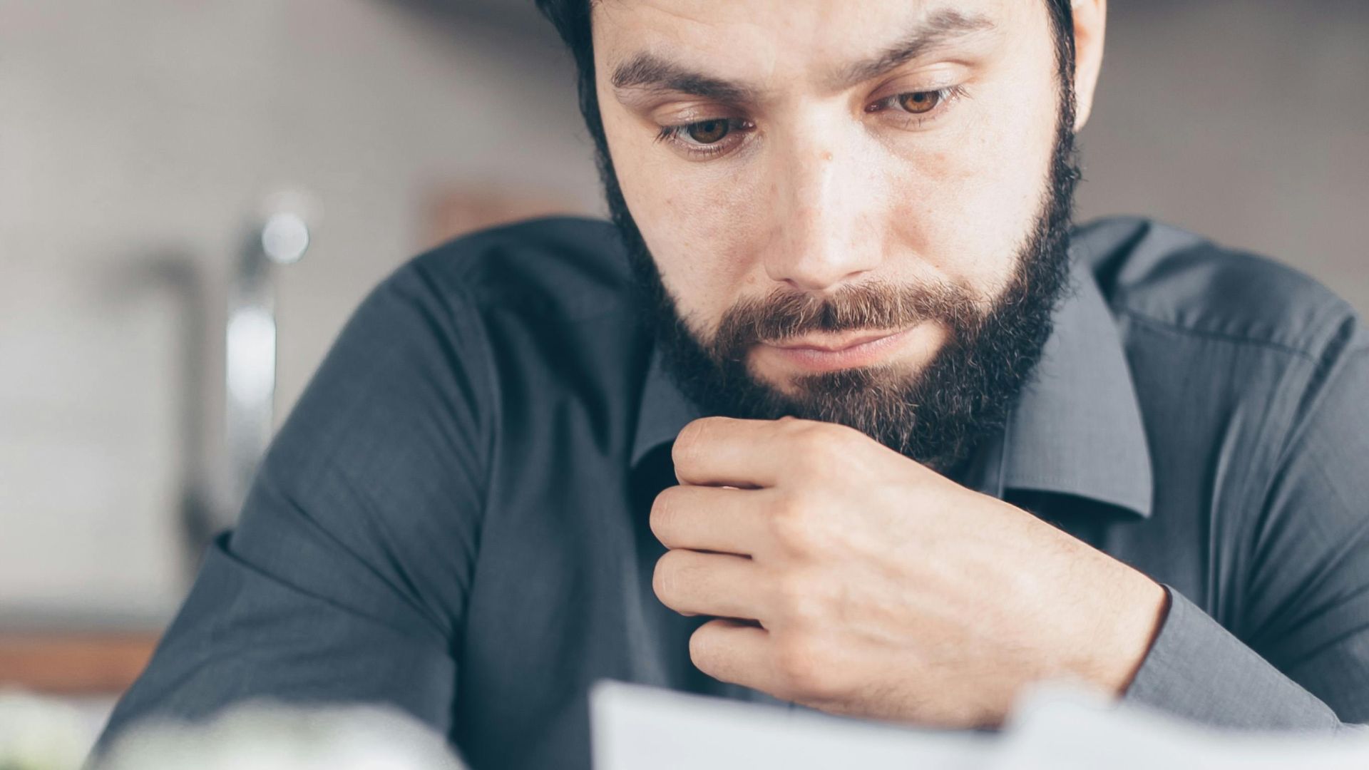 A bearded man in a gray shirt intently reads documents while seated indoors, appearing concerned.