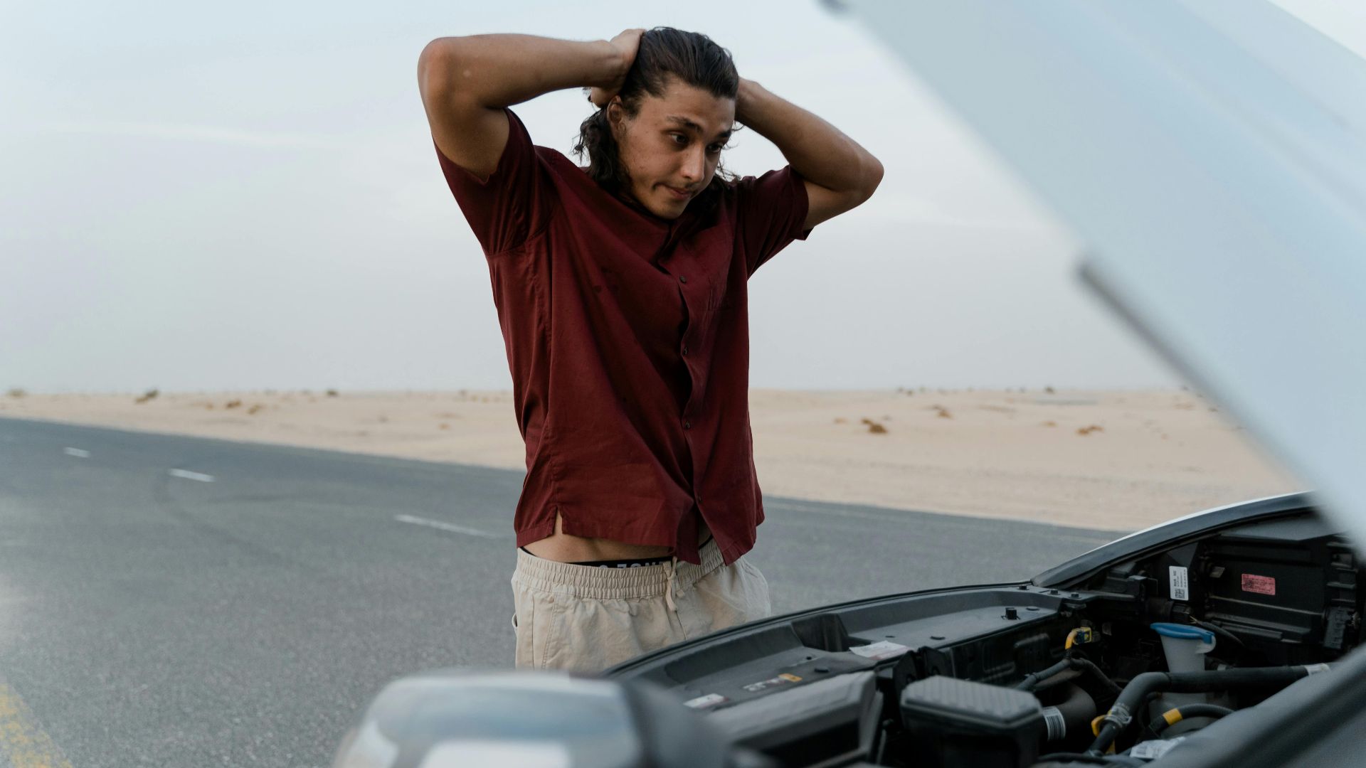 A frustrated man examining a broken car engine on a deserted road, showing stress and concern.