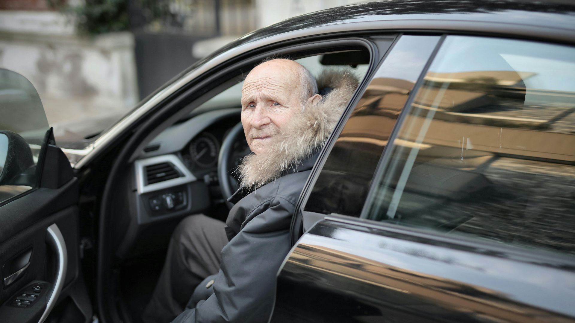 An elderly man in a winter coat stepping out of a parked car outdoors.