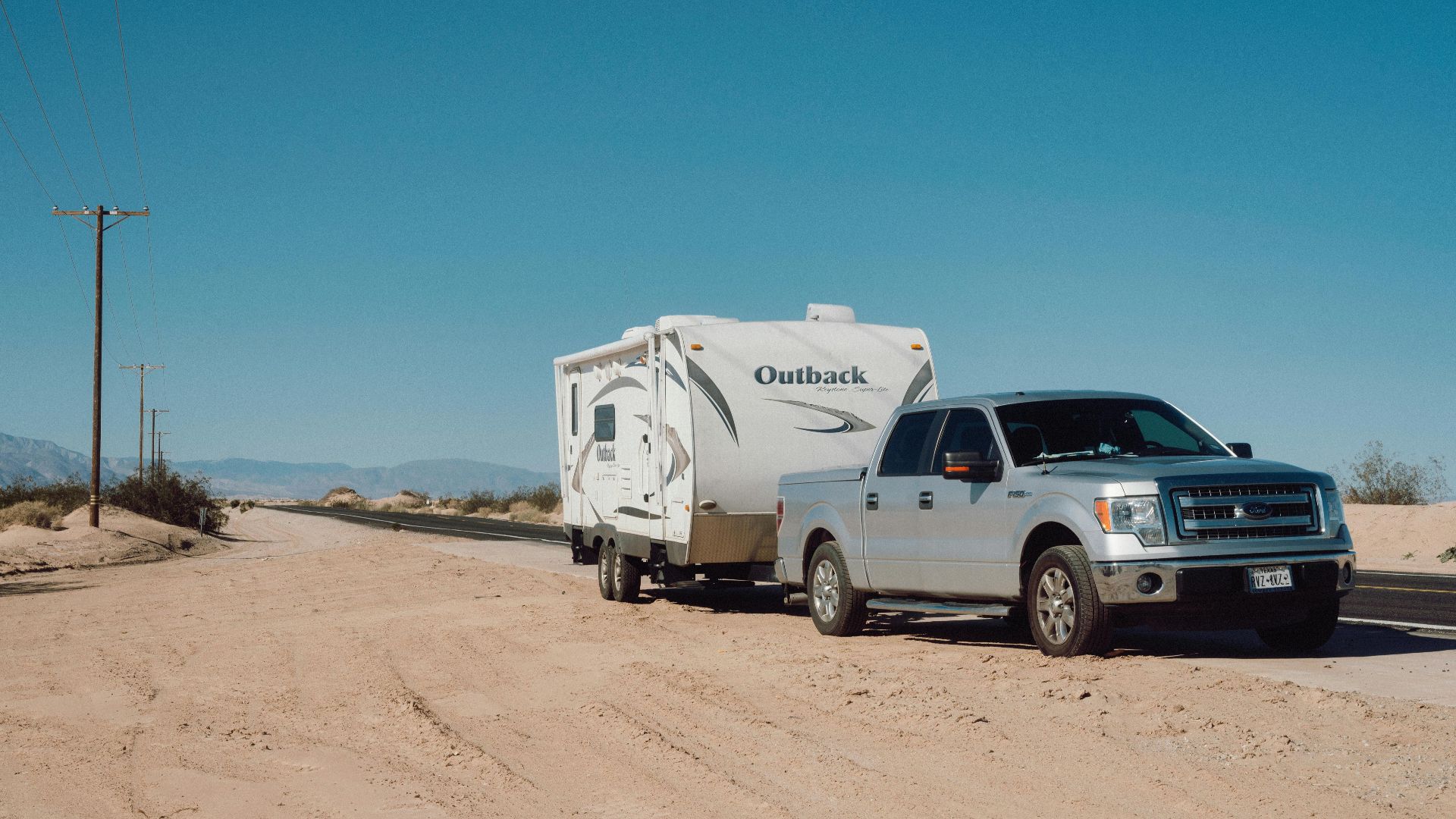 Silver truck towing an Outback trailer on a desert road under clear blue skies.