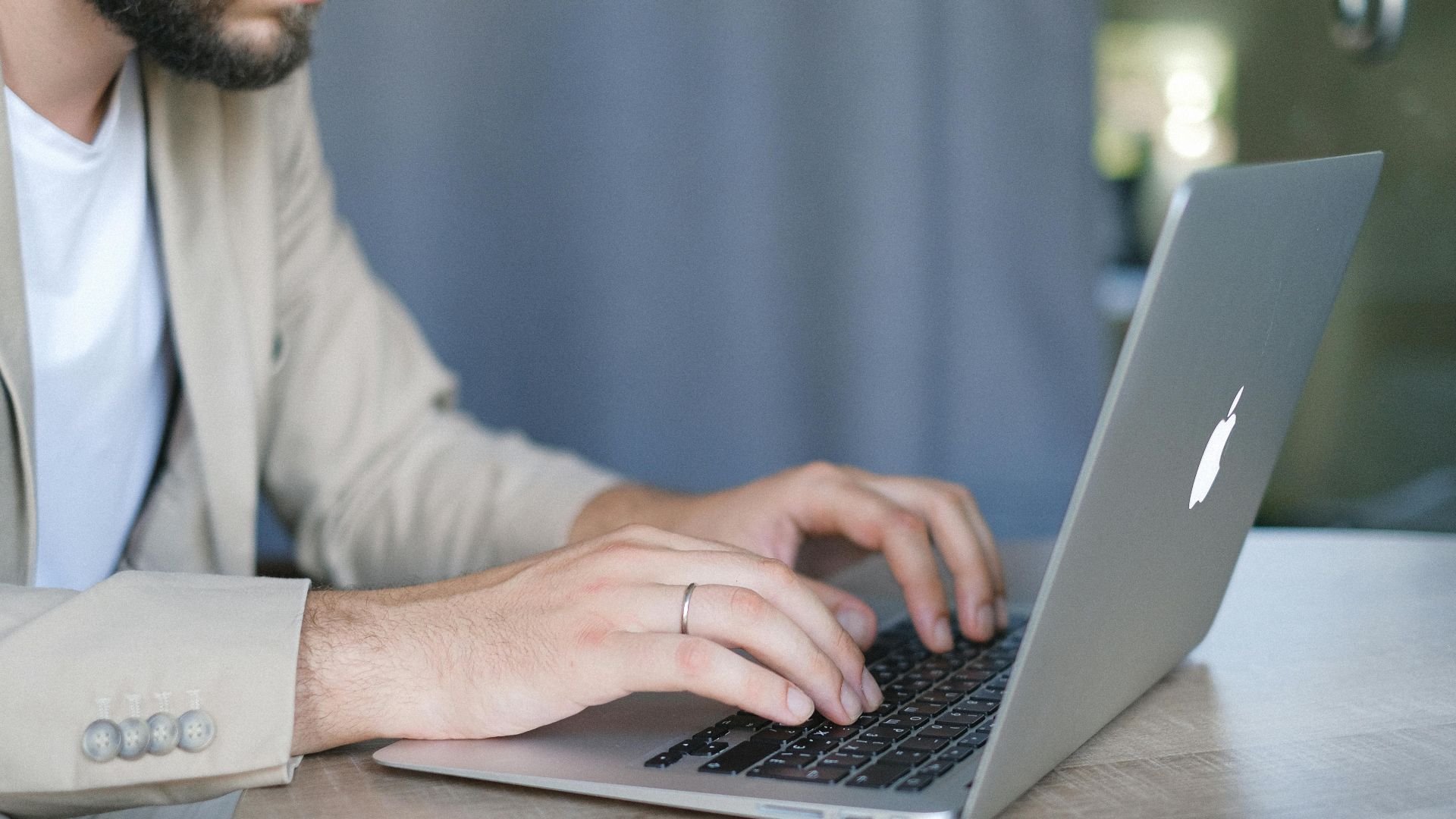 Side view of crop bearded male entrepreneur wearing classy jacket sitting at wooden table and browsing modern netbook in daylight