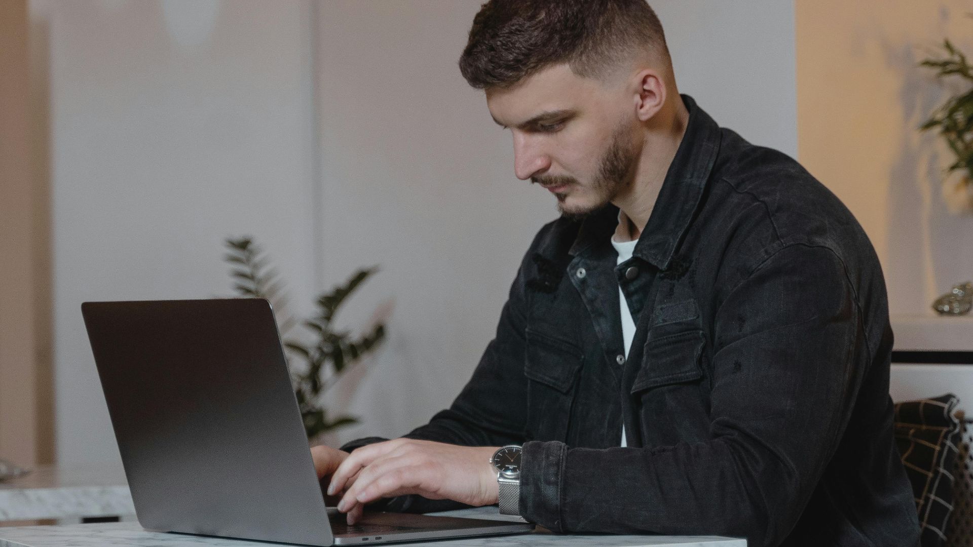 Young man in a denim jacket working on a laptop indoors, showcasing modern remote work lifestyle.