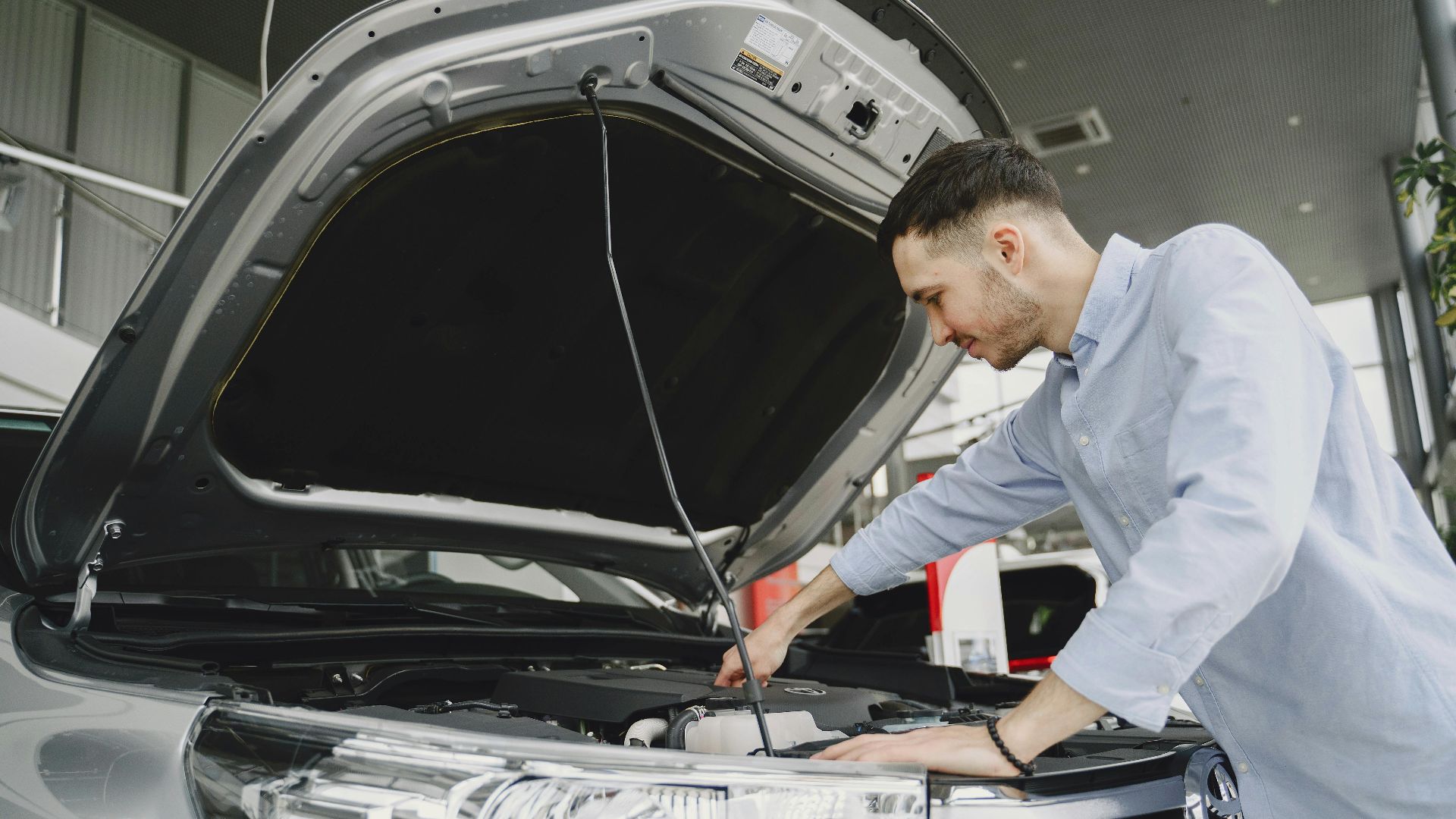 A man in a blue shirt examines a car's engine in a modern vehicle showroom.