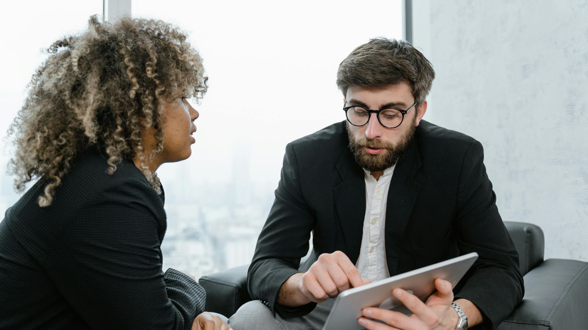 A focused business meeting between two professionals discussing data on a tablet indoors.