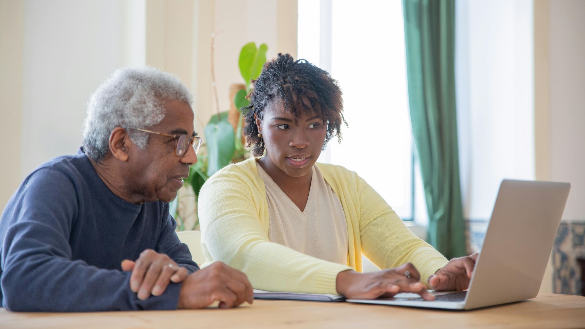 Young woman assists an elderly man with laptop indoors, highlighting intergenerational tech support.