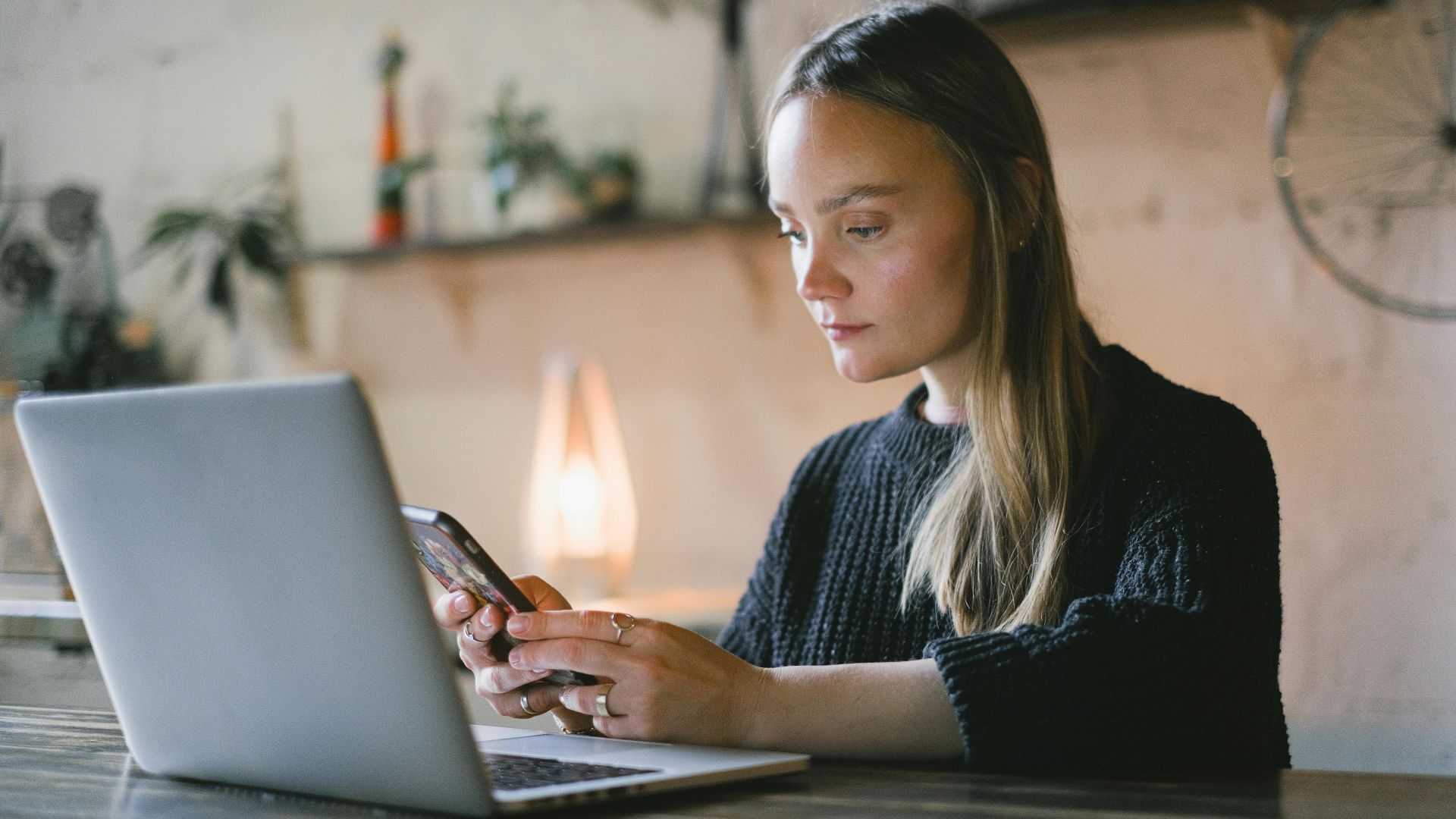 Concentrated female freelancer text messaging on cellphone while sitting at table with netbook during online work on blurred background