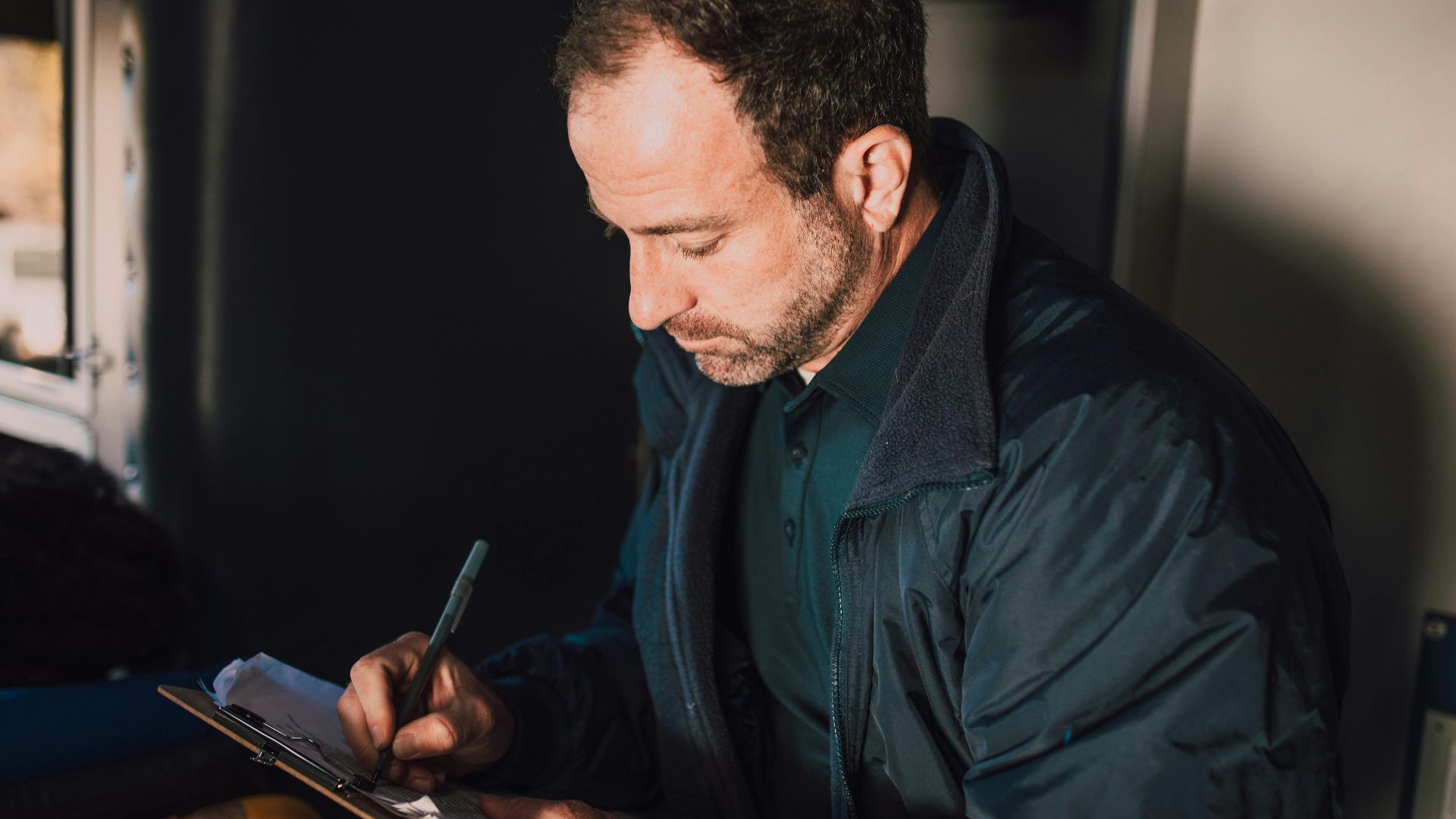 Adult male writing notes on a clipboard inside. Concentrated and thoughtful atmosphere.