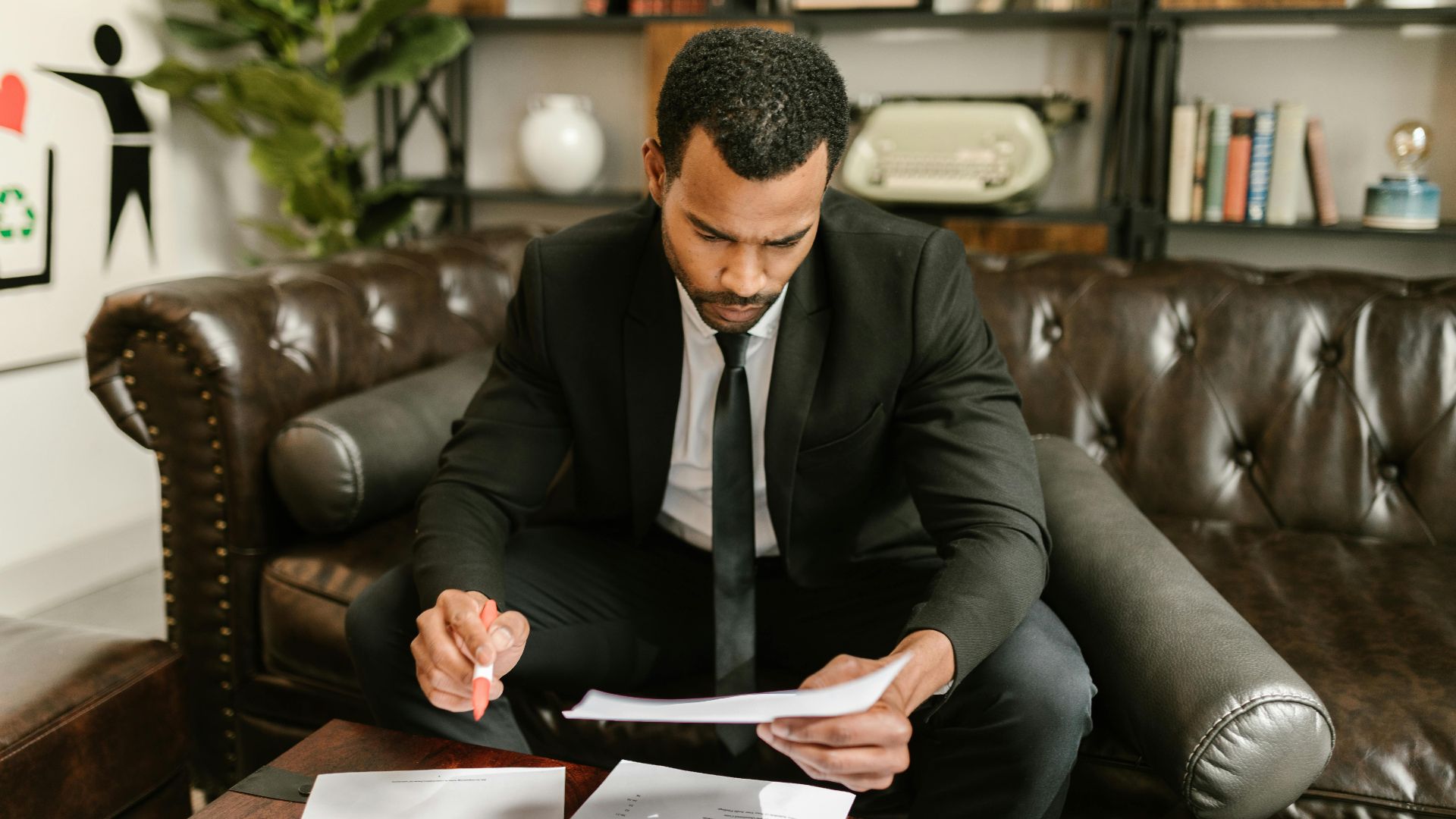 A businessman in a suit attentively reviews paperwork on a brown leather sofa.