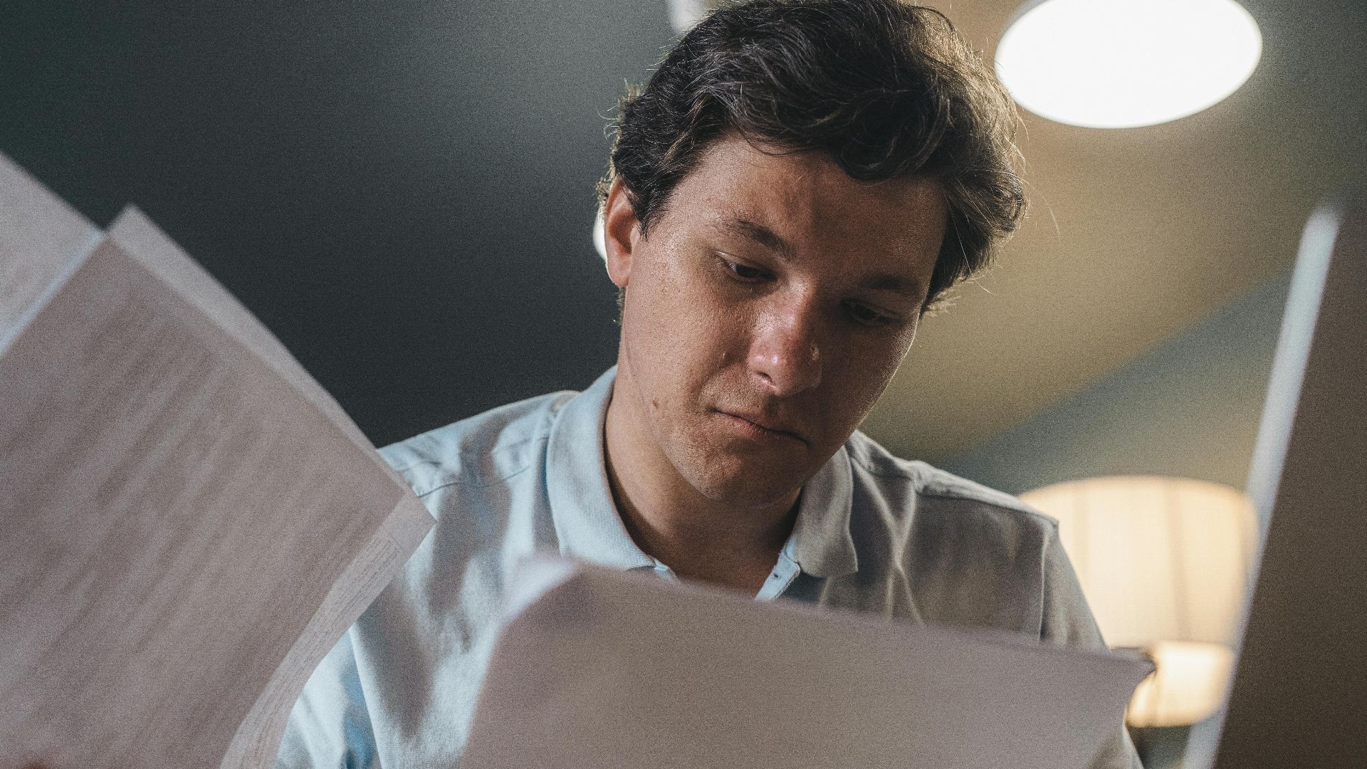 Serious man wearing a light blue shirt intently reading printed papers indoors under warm lighting.