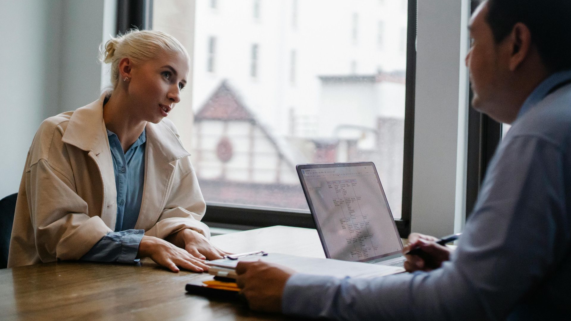 Crop man sitting at wooden desk with laptop and documents and listening attentively to positive young woman in trendy outfit in light workspace