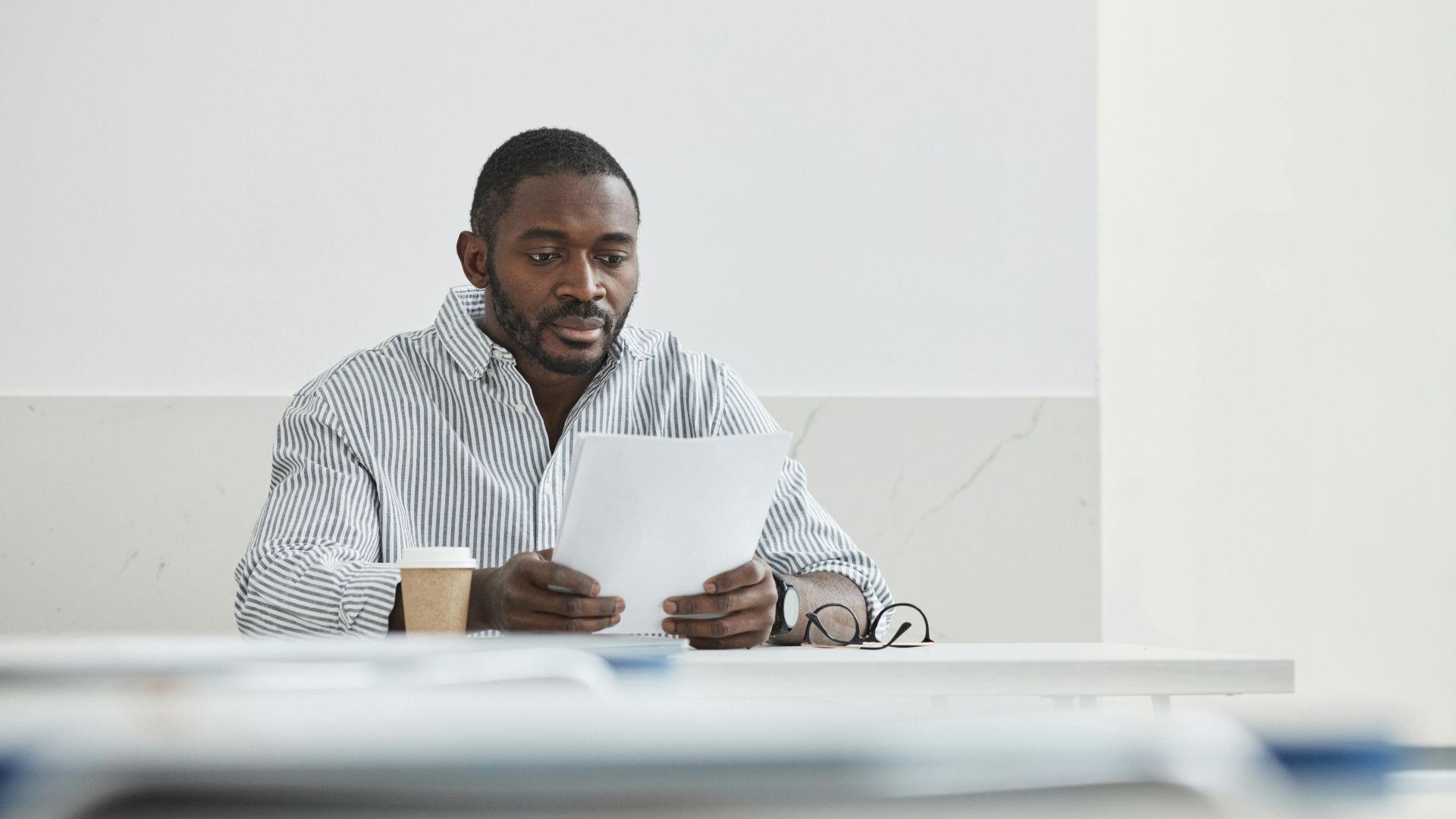African American man sitting indoors, reading papers with a coffee cup nearby.