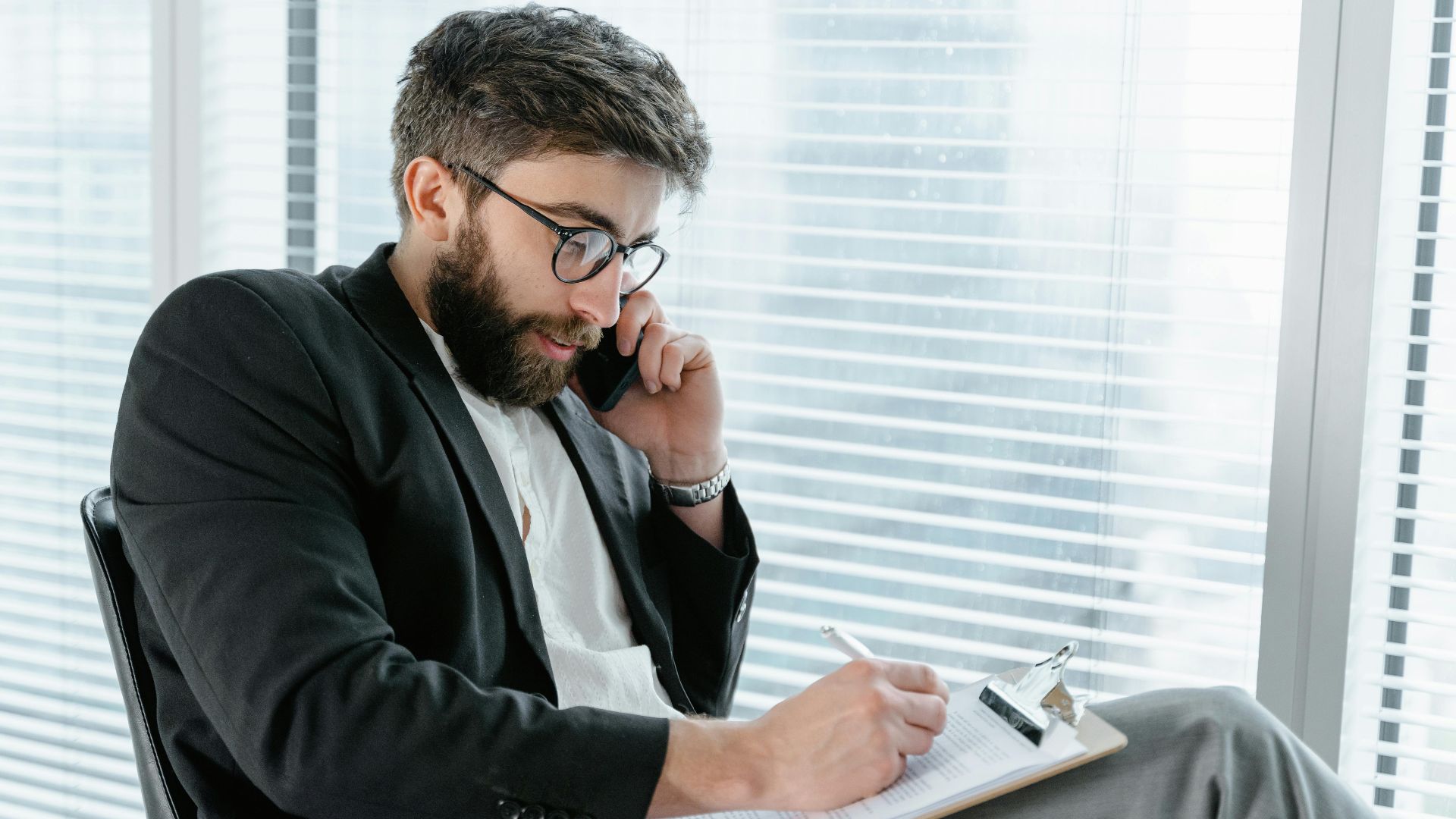 A professional man in a suit writing on a clipboard while talking on a smartphone in an office.