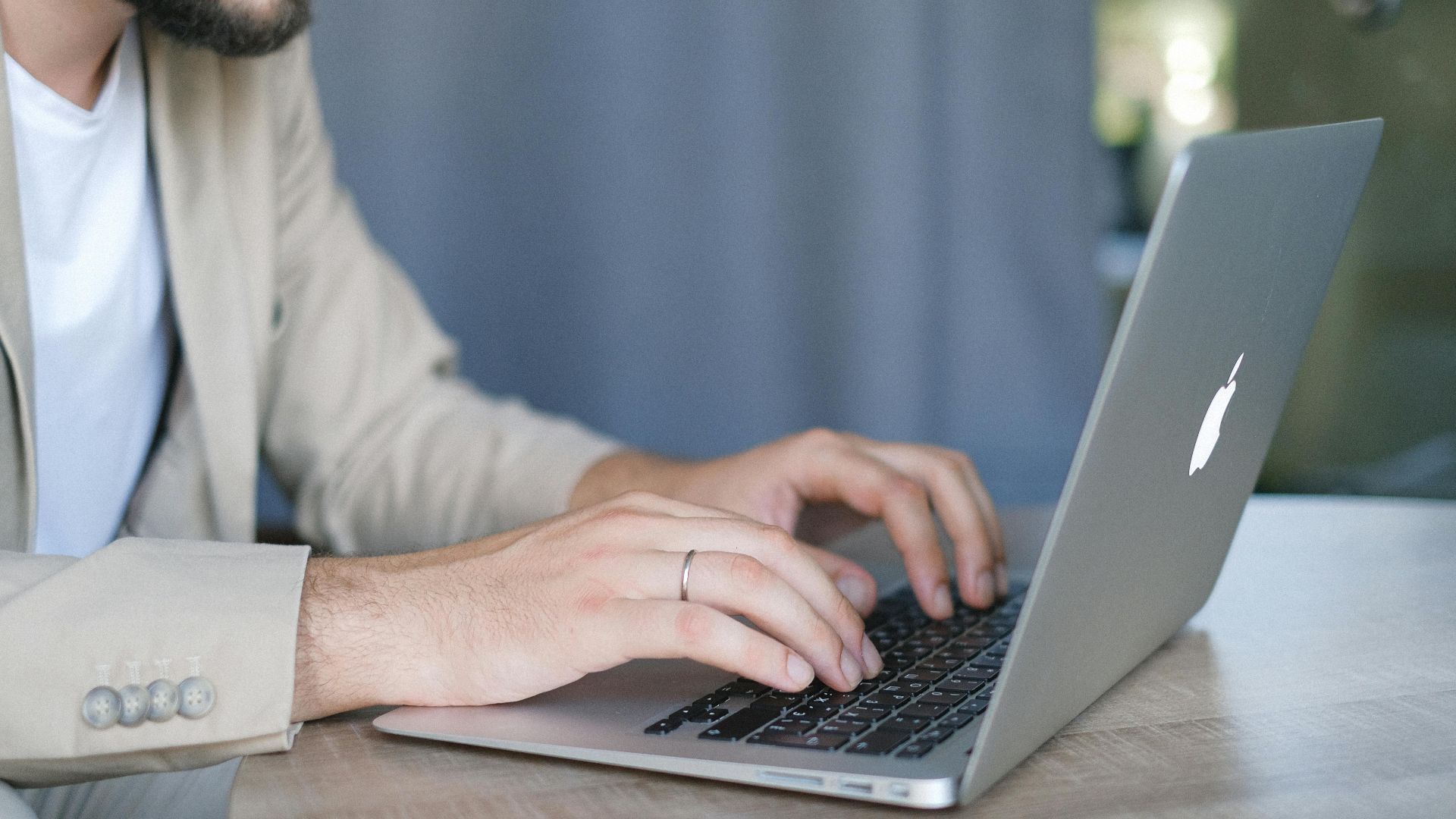 Side view of crop bearded male entrepreneur wearing classy jacket sitting at wooden table and browsing modern netbook in daylight