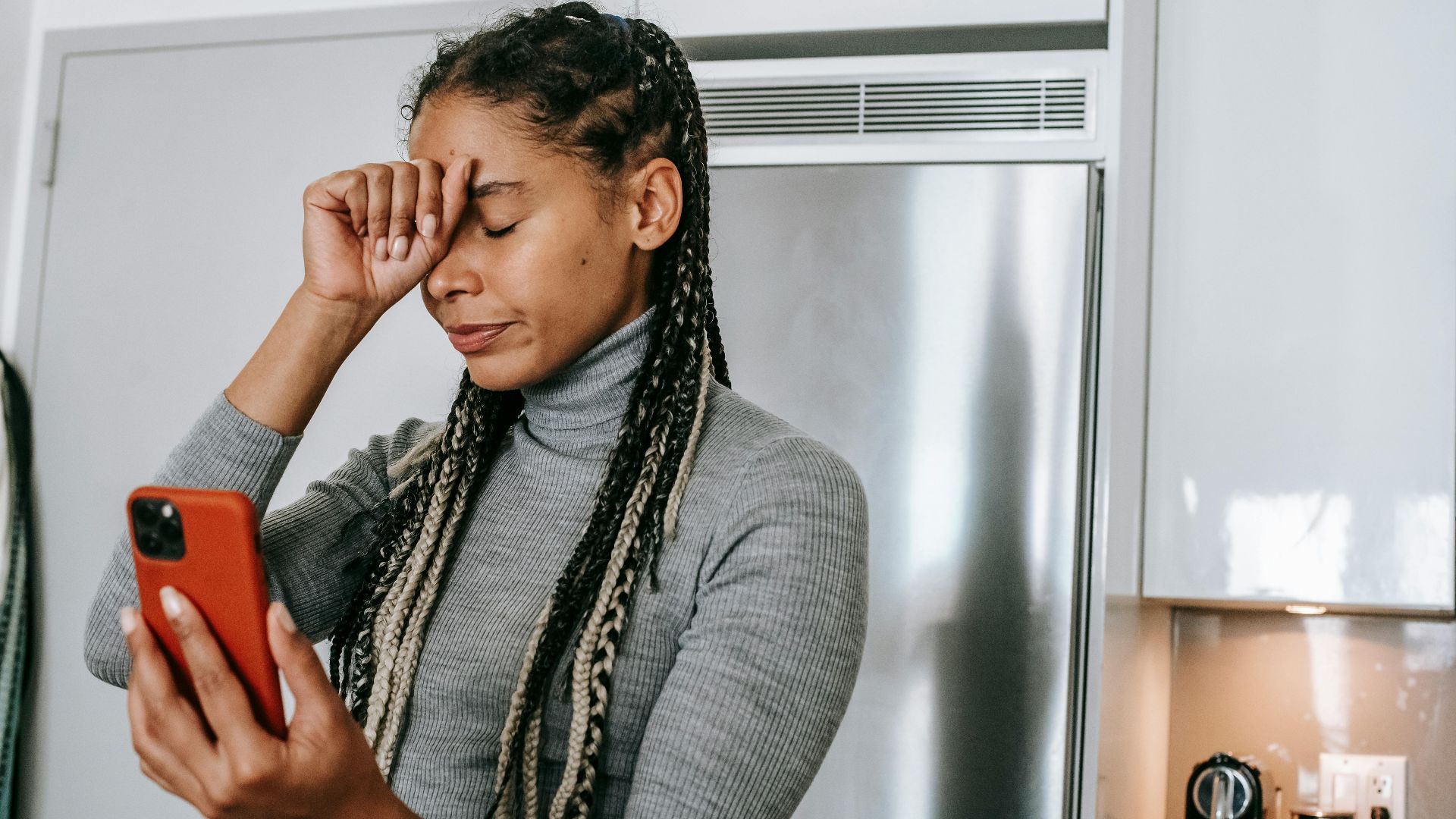 Unhappy young African American female with long braids in turtleneck touching face with closed eyes while having video conversation on smartphone in kitchen