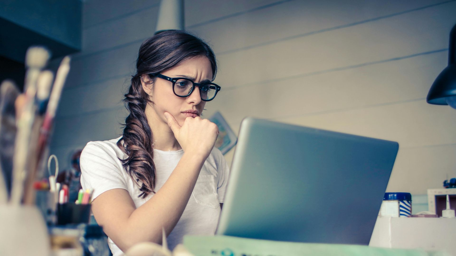 Young woman with glasses deeply focused on a laptop surrounded by art supplies in a home office.