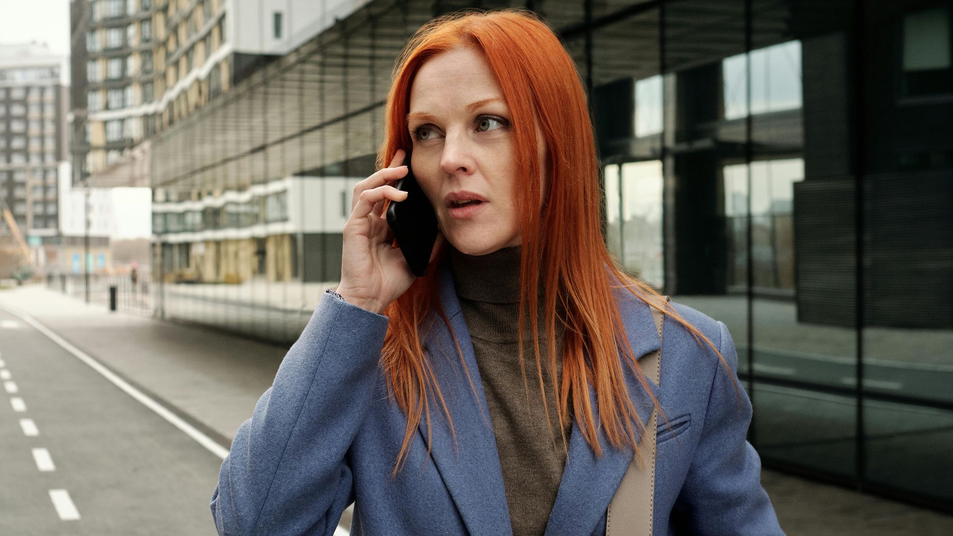 Red-haired woman in blue coat making a phone call on an empty city street.