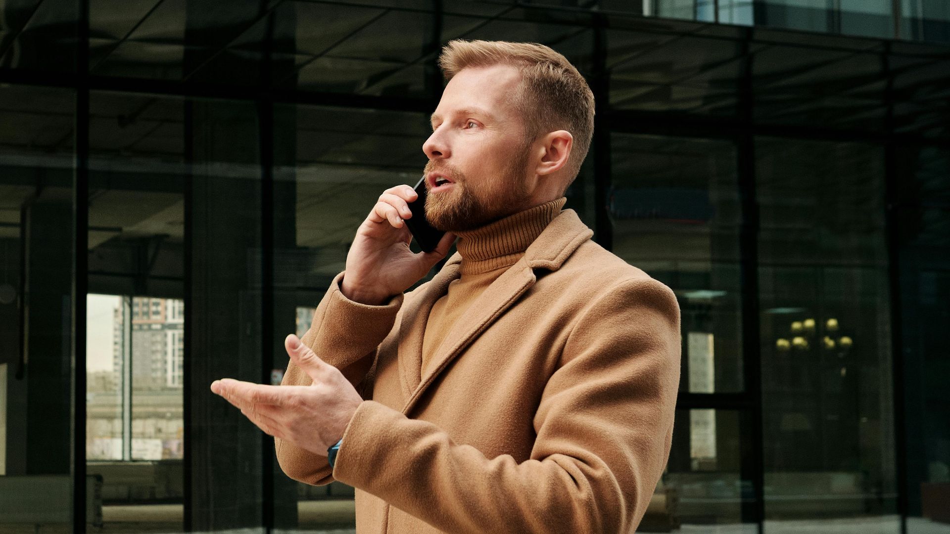 Confident businessman in a brown coat talks on phone outdoors in city.