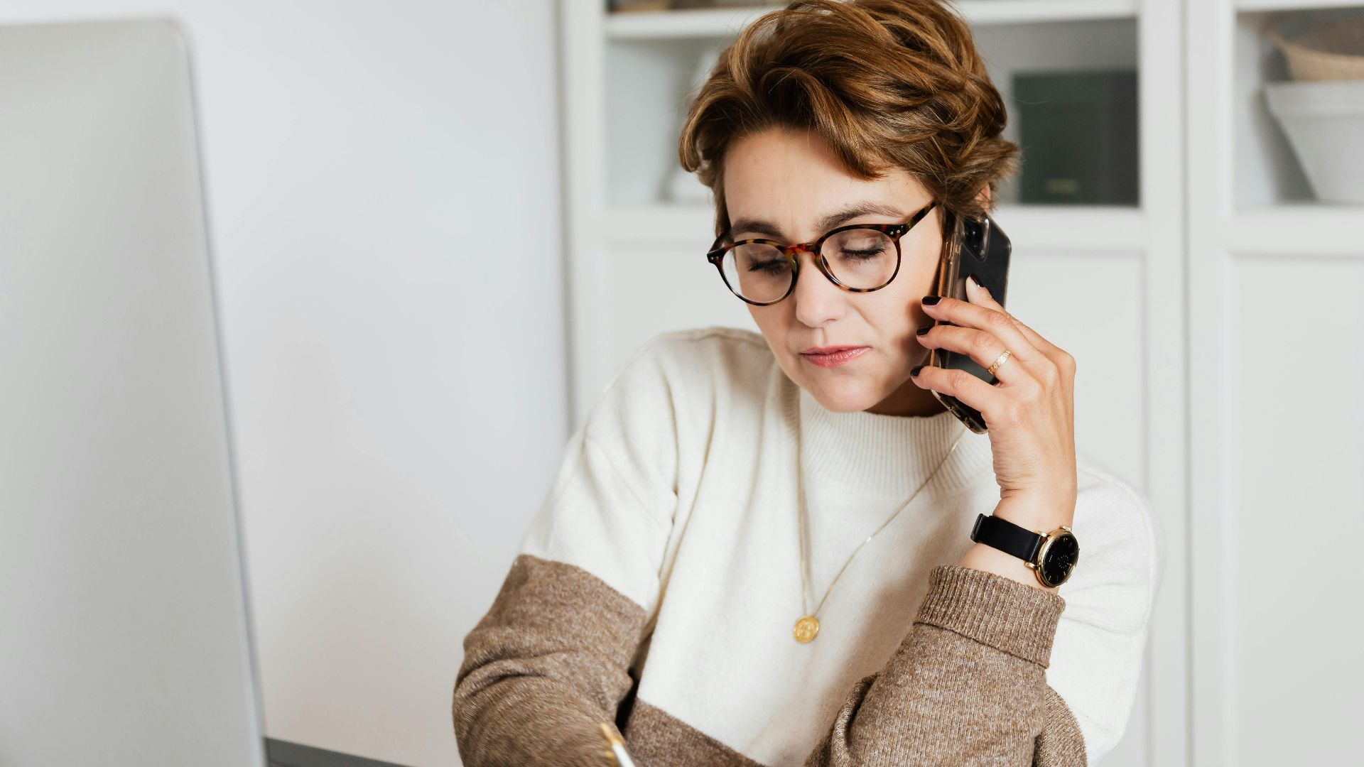Good looking short haired female worker listening to work instructions on mobile and noting down information in modern office