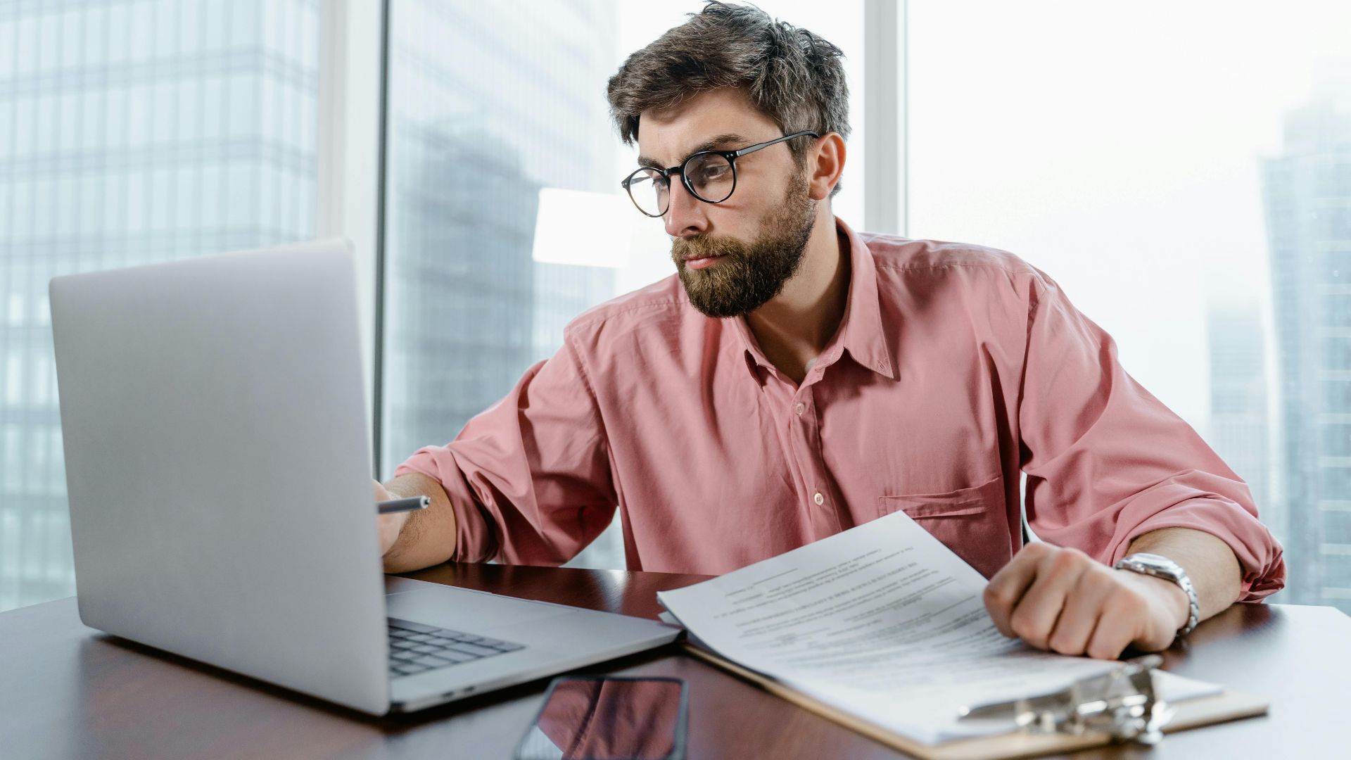 A businessman in an office reviews documents and data on a laptop, concentrating on financial analysis.