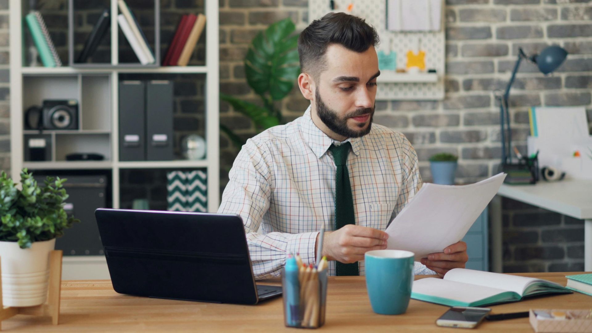 Young man reading papers at desk with laptop and office decor, focused on work.