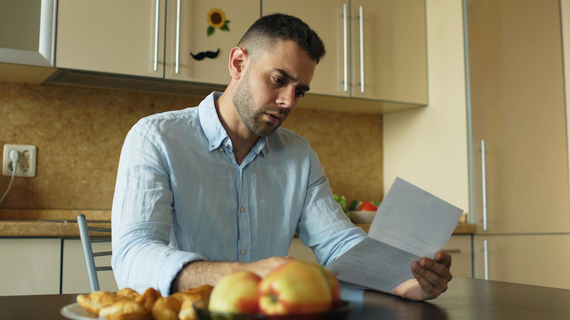 Man in a kitchen reading a document, apples on table.