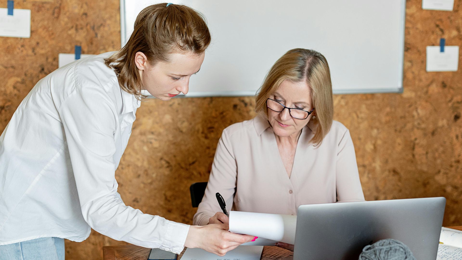Two women discussing work documents in a modern office setting with a laptop.