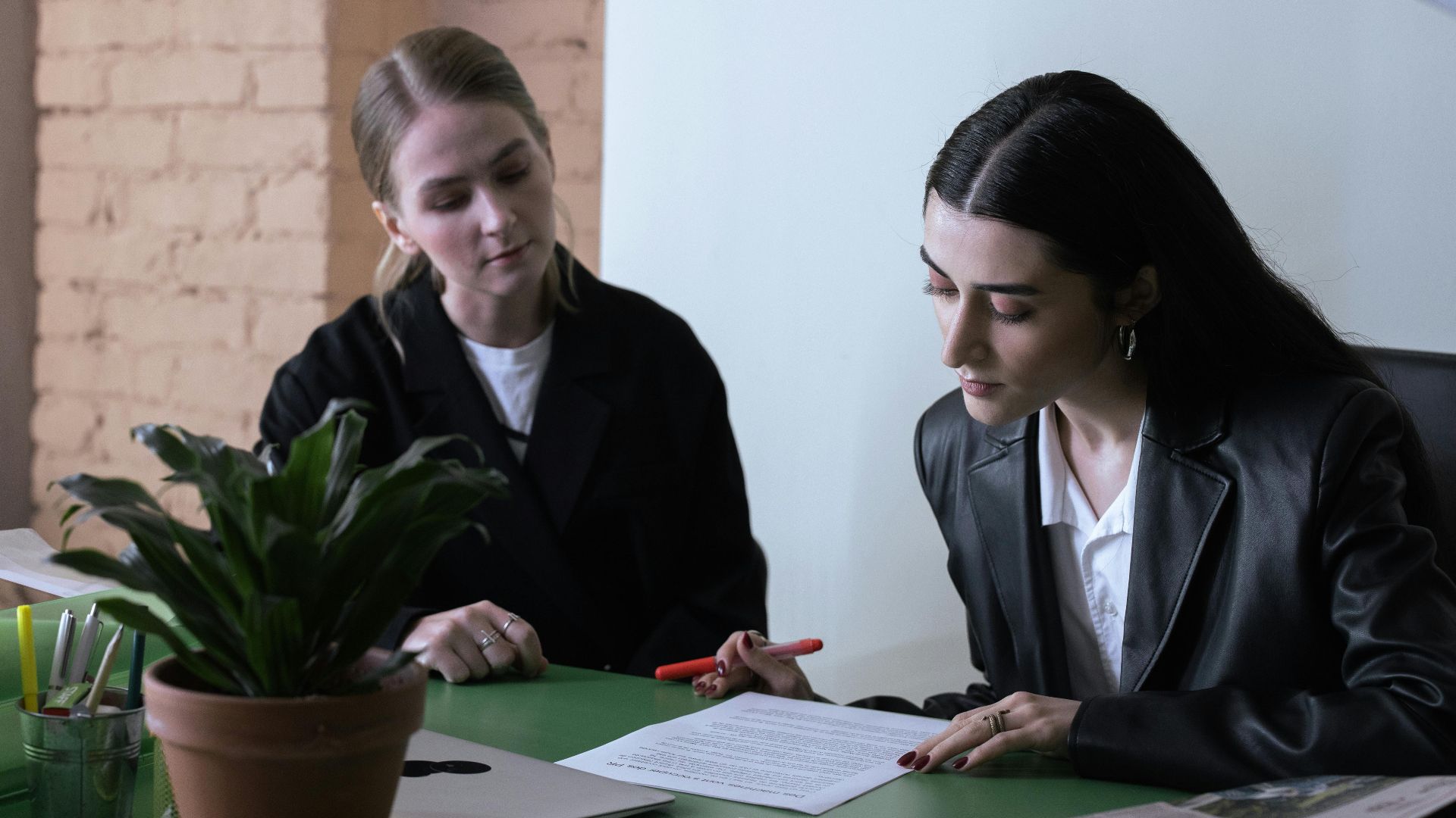 Two women collaborate in a modern office, reviewing documents at a desk.