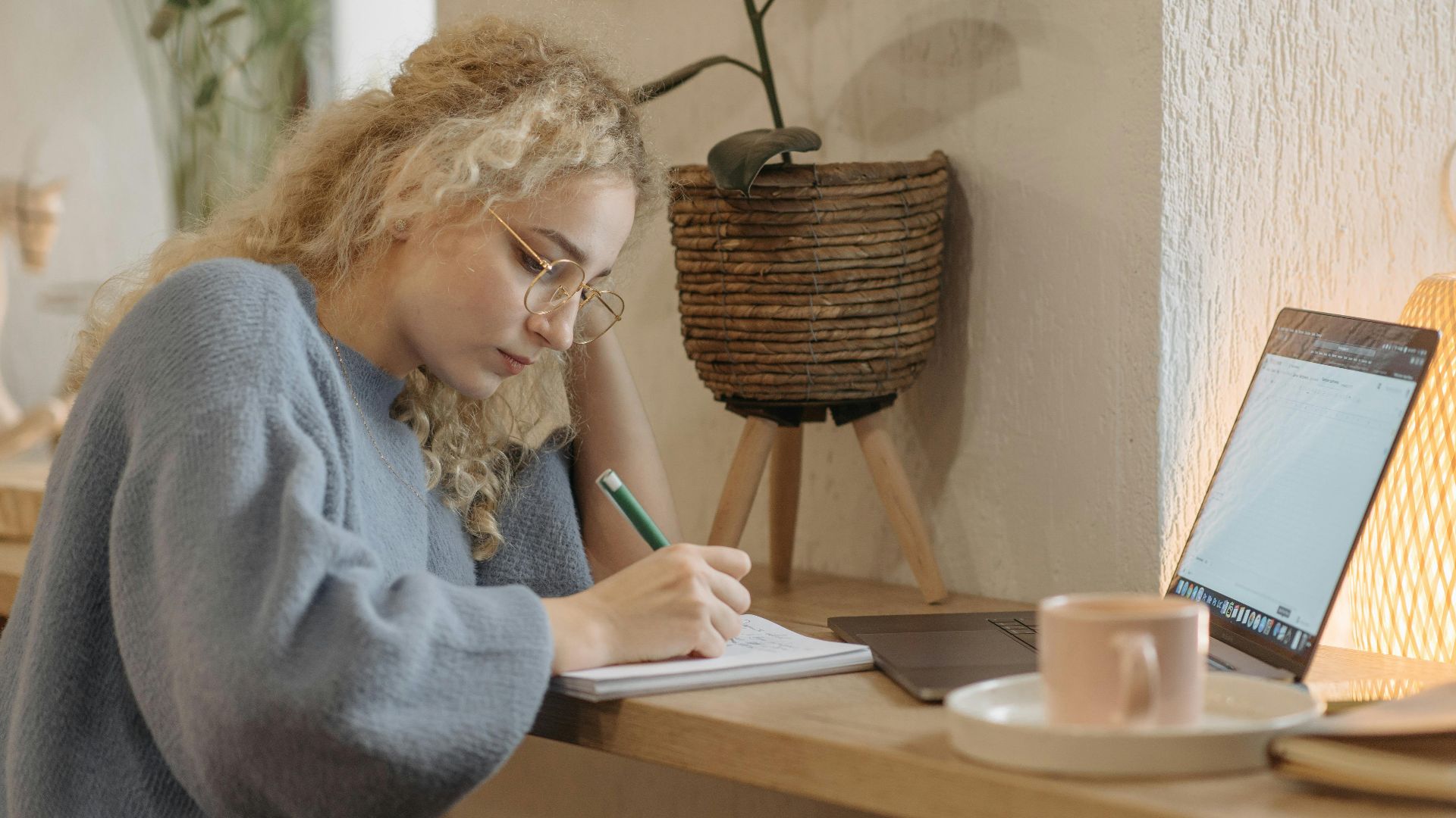 Young woman writing in notebook at cozy desk with laptop and coffee.