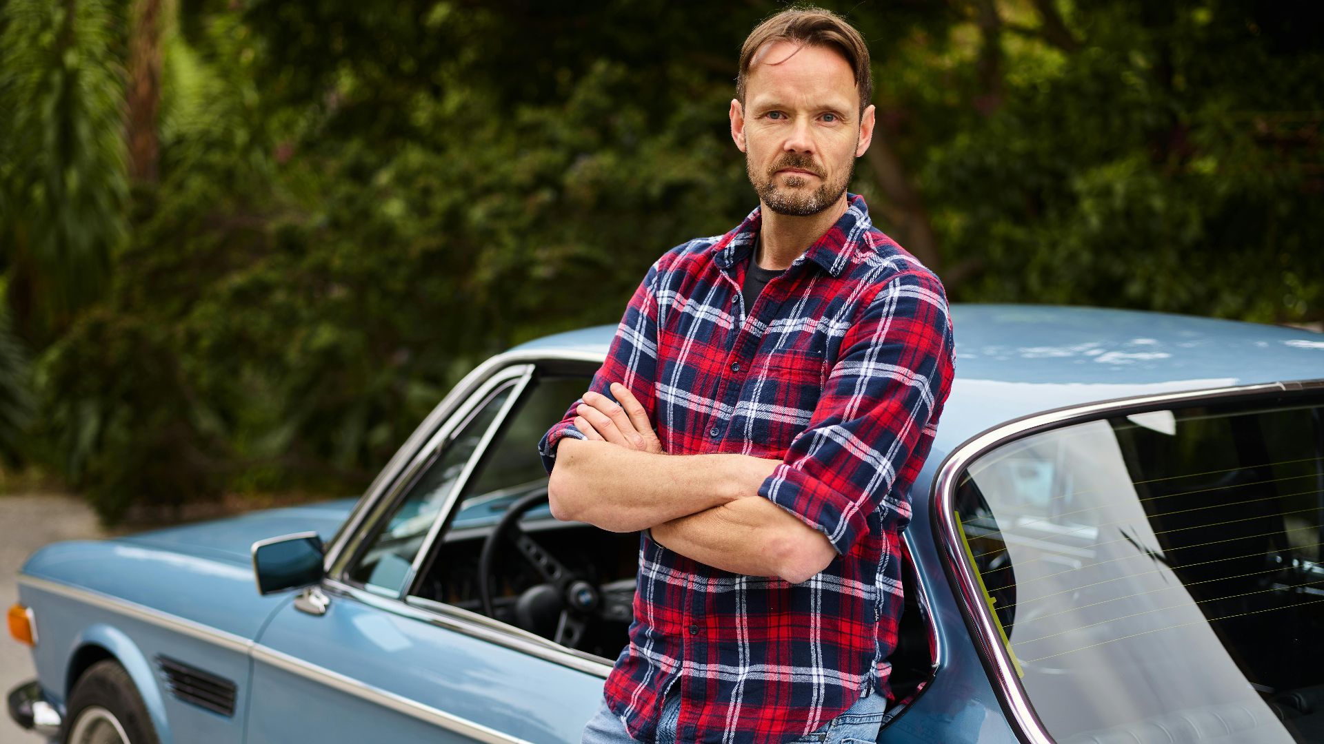 Adult bearded male in jeans and shirt standing with crossed arms leaned on blue retro auto near forest in daylight and looking at camera