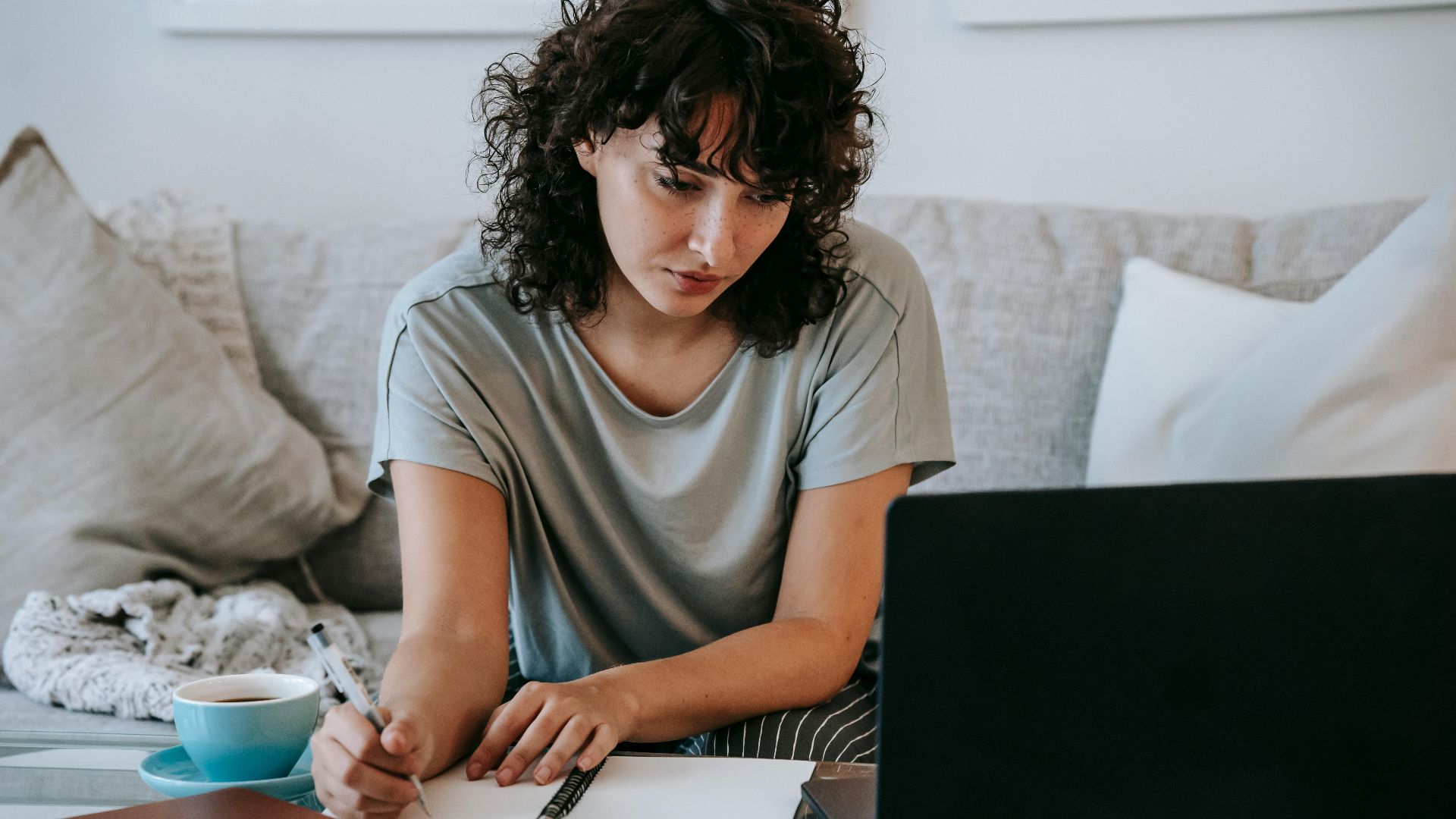 Focused young female student with dark curly hair in casual clothes taking notes in copybook while preparing for exams sitting on couch and using laptop at home
