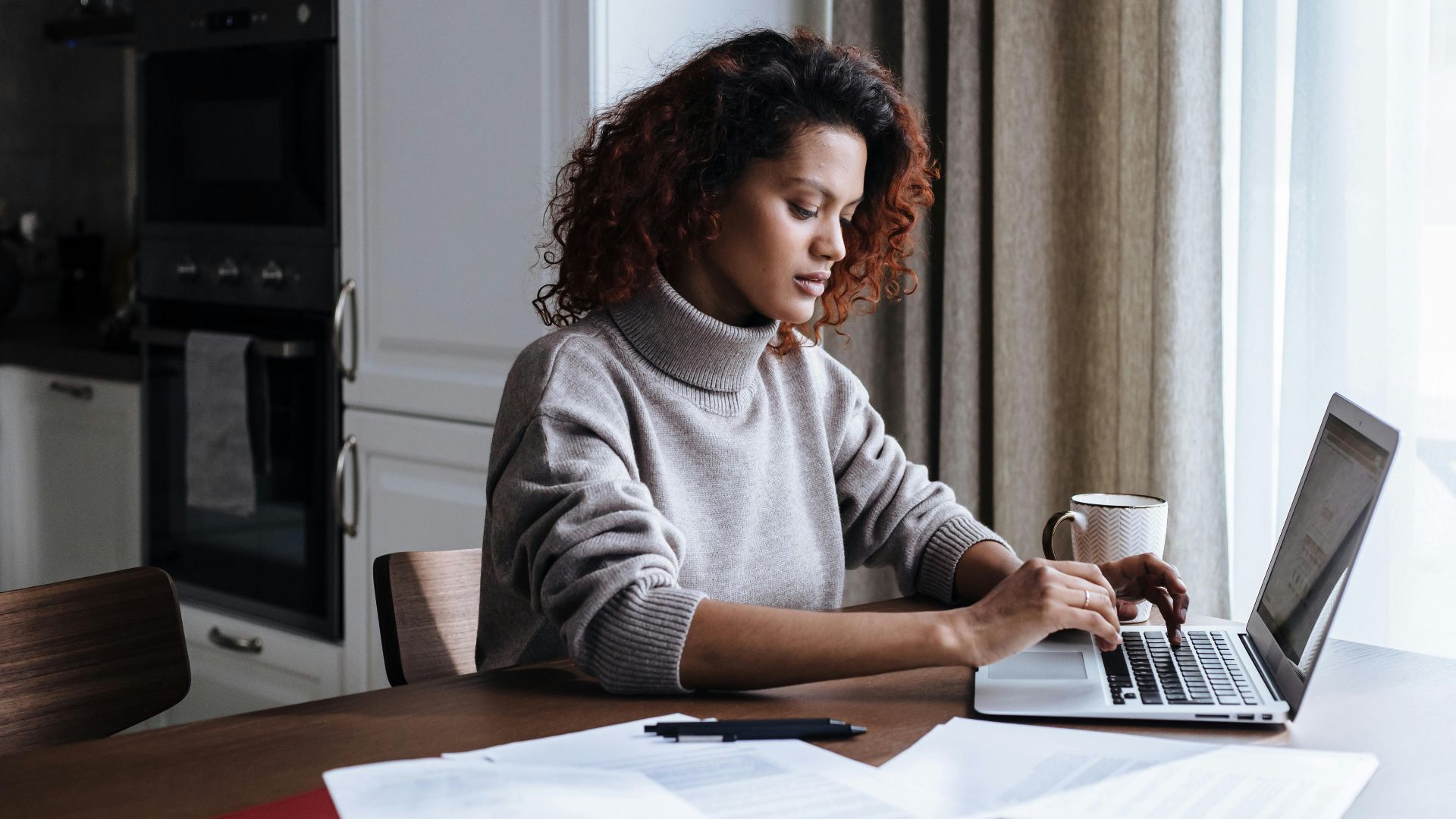 Focused young woman working on laptop at kitchen table with documents.