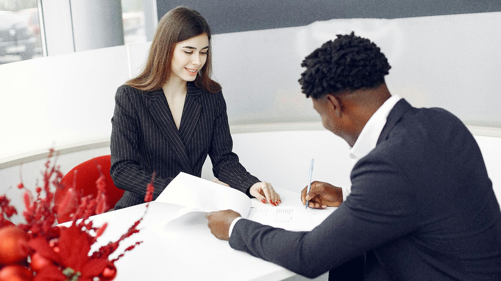 Positive stylish multiethnic buyer in formal classy suit and agent signing papers in modern light creative office in daylight and smiling happily