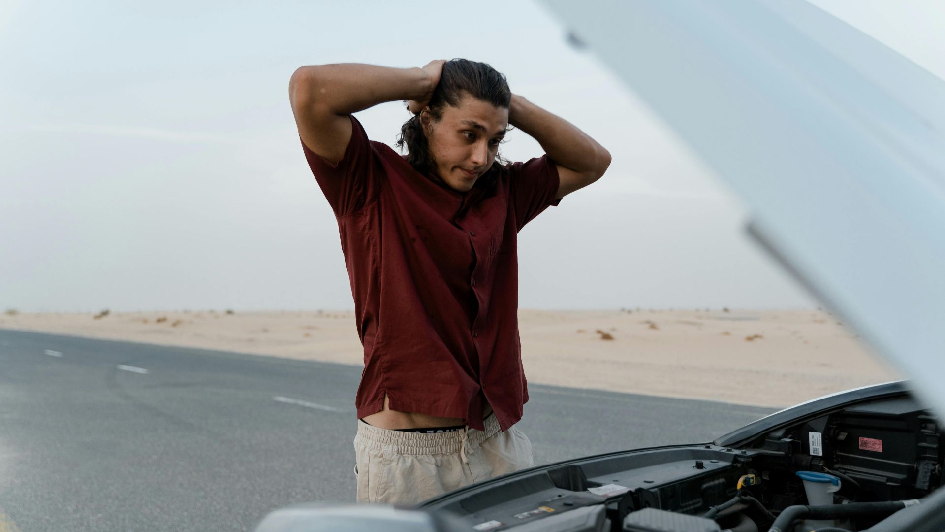 A frustrated man examining a broken car engine on a deserted road, showing stress and concern.