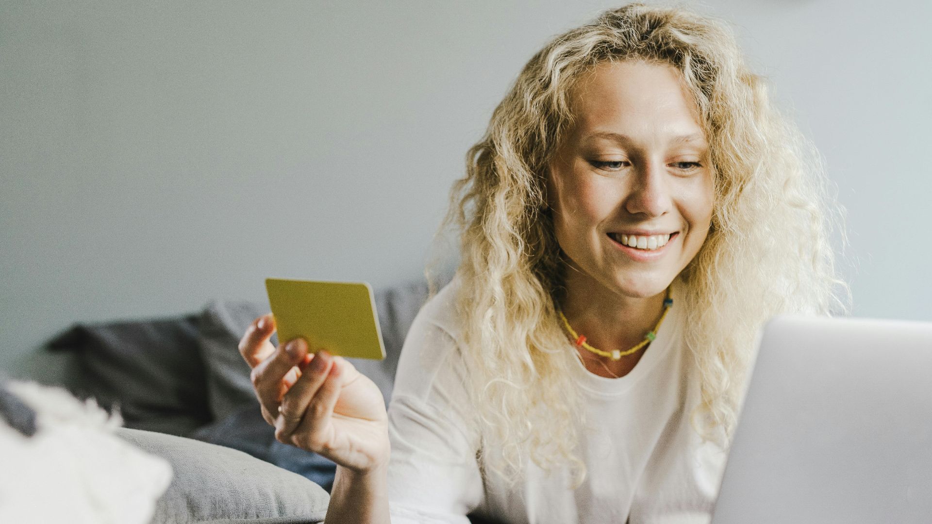 Smiling blonde woman with curly hair shopping online with a bank card indoors.