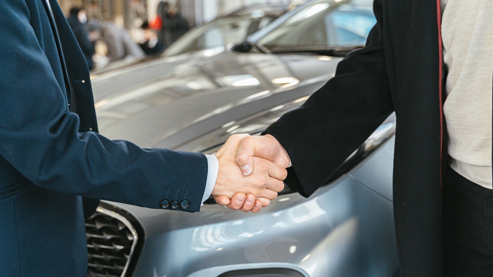 A close-up of two people shaking hands in front of a car in an indoor setting.