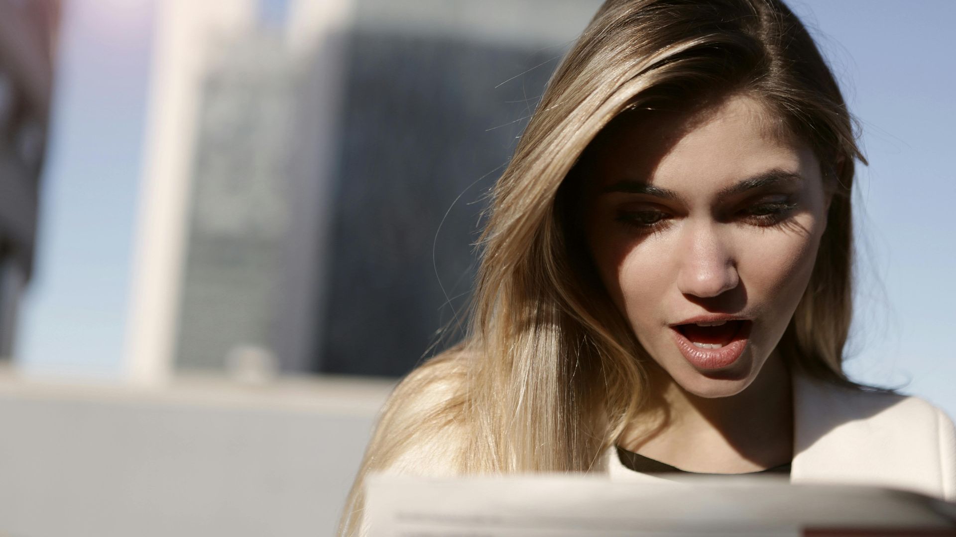 A young woman with blonde hair expresses surprise while reading a newspaper outdoors in daylight.