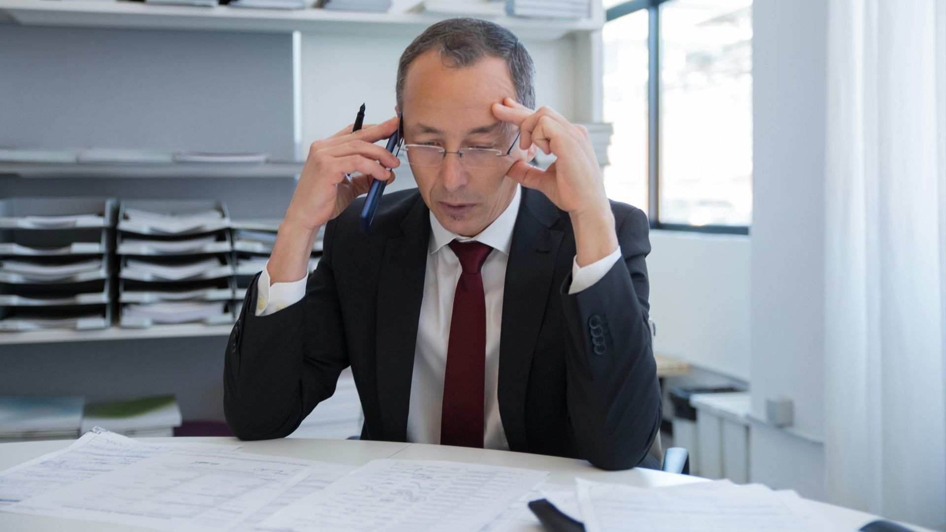 Businessman in a black suit stressed over documents in a bright office setting.