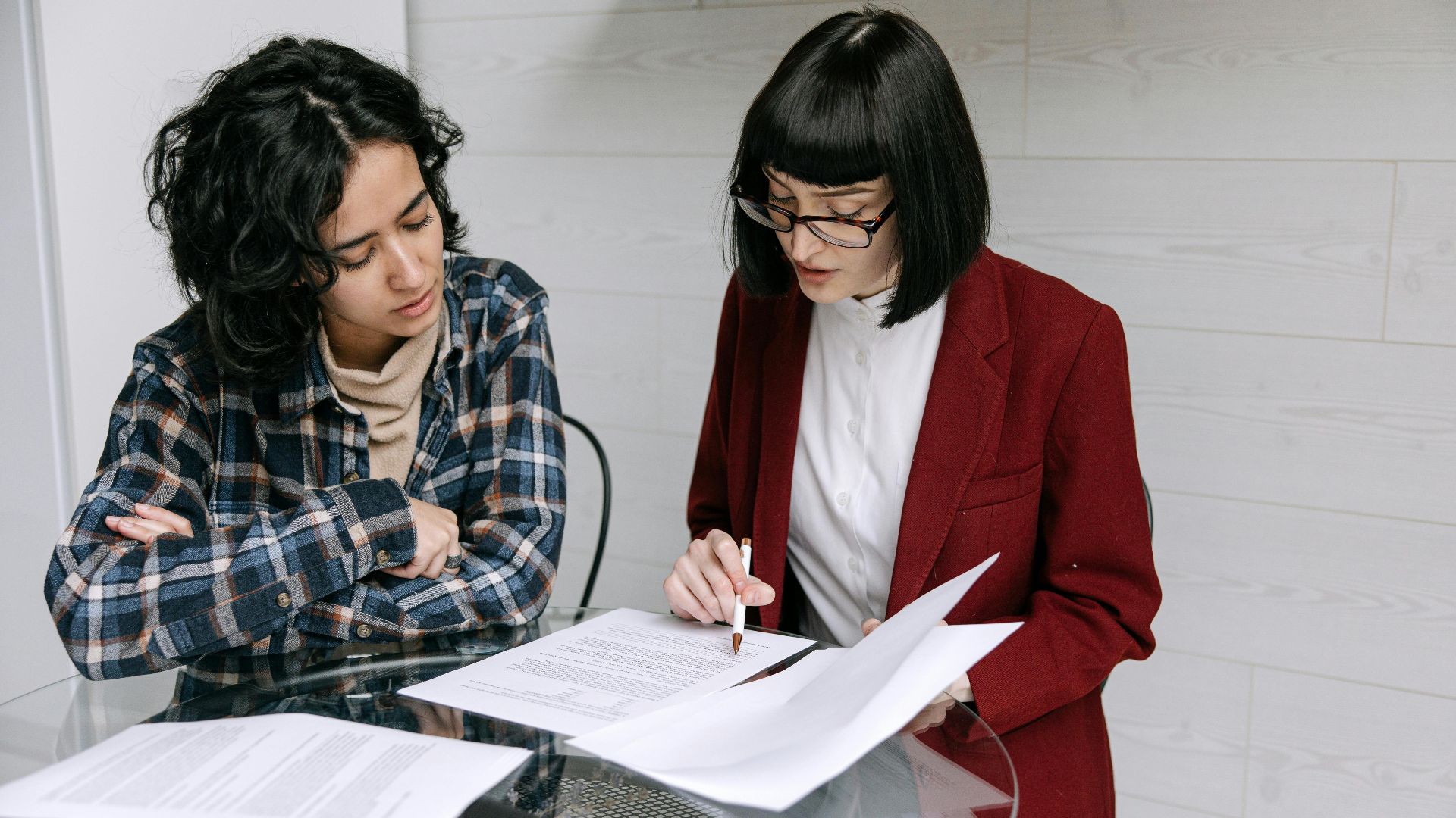 Two women collaborating at a glass table, reviewing business documents in a modern office setting.