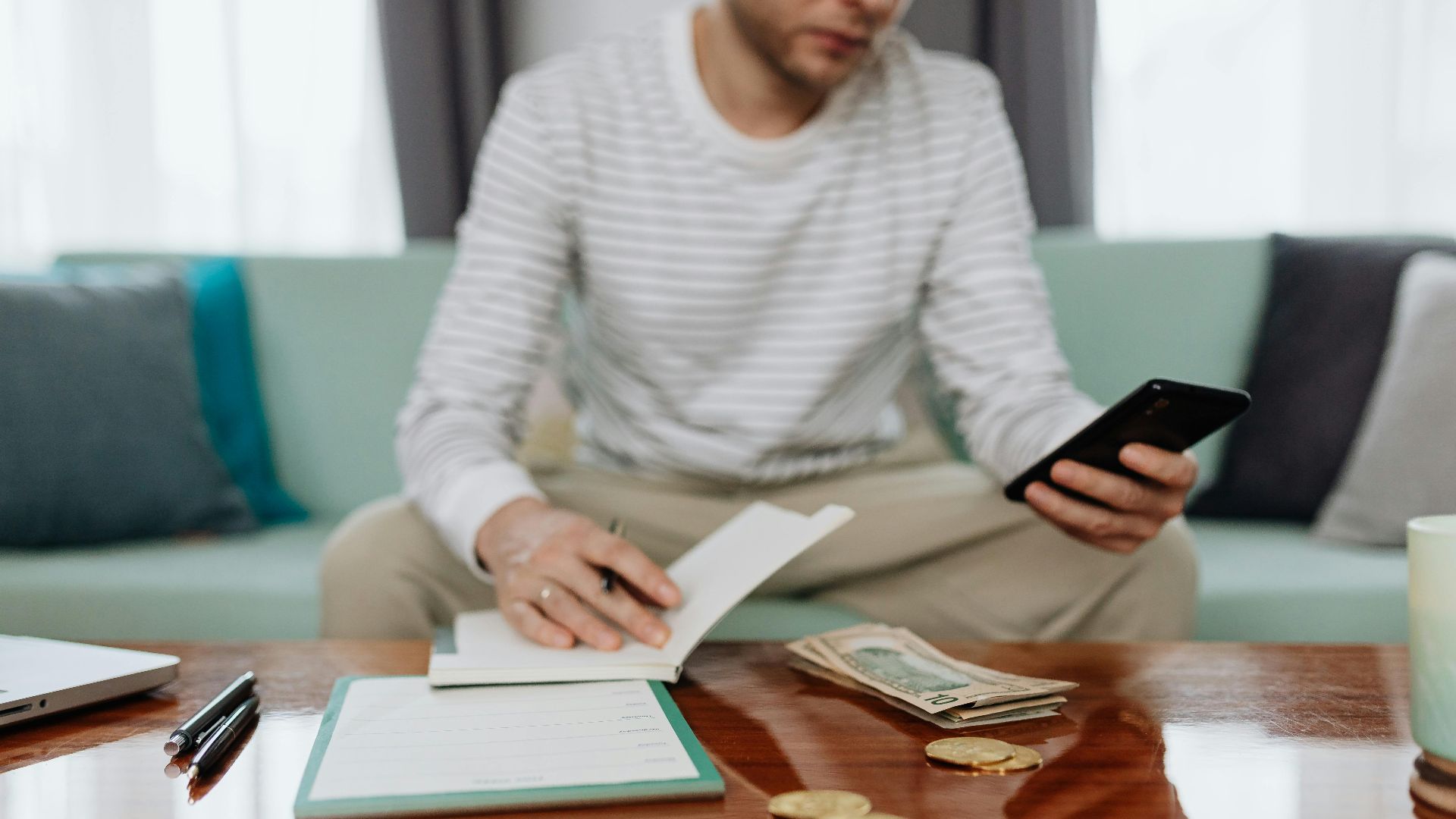 Adult man sits on sofa managing finances with cash, smartphone, and notebook.