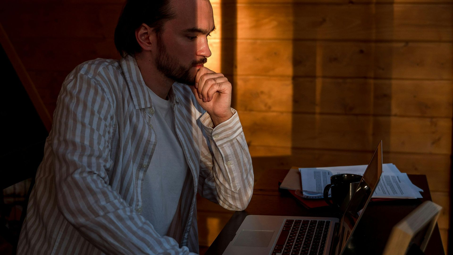 A man is deeply concentrating on his laptop while seated at a desk, basked in warm sunlight