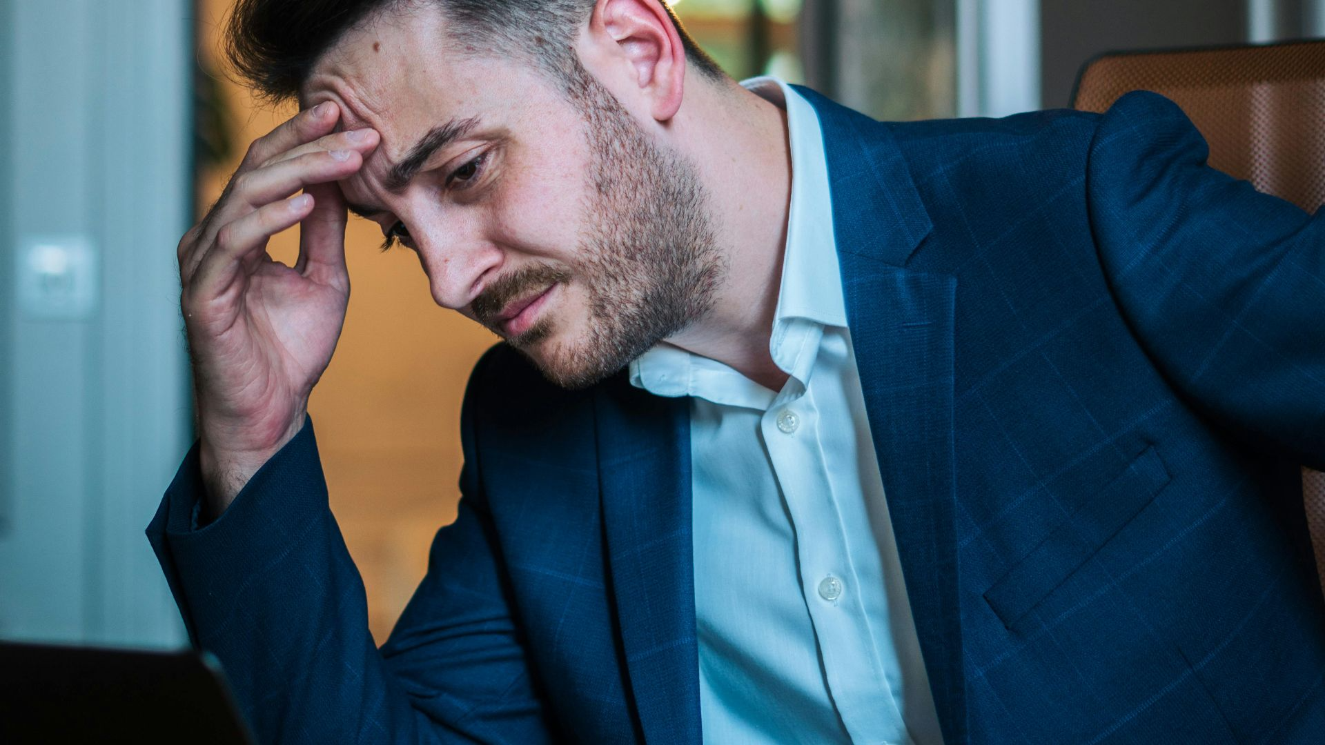 A worried businessman in a blue suit working on his laptop in an office environment