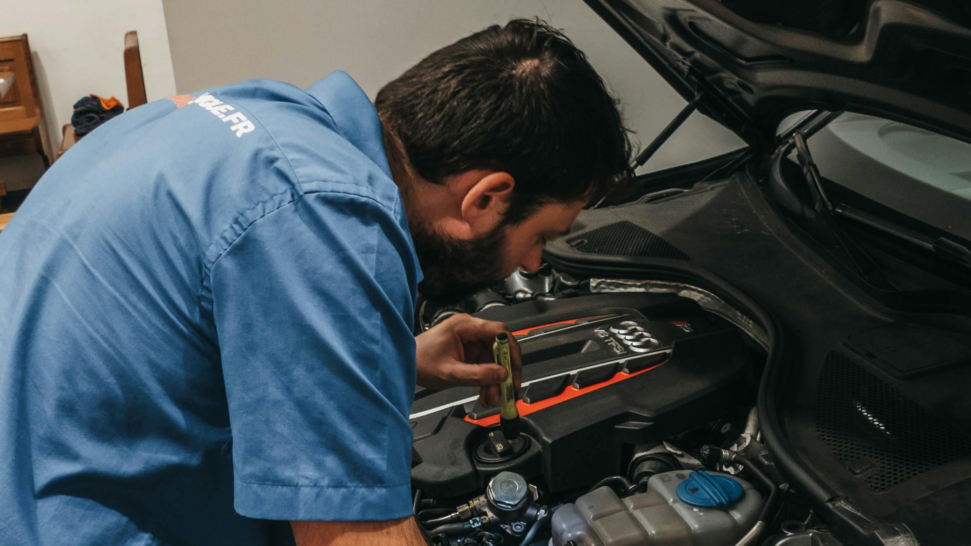 Mechanic in a workshop examining a car's engine under an open hood.