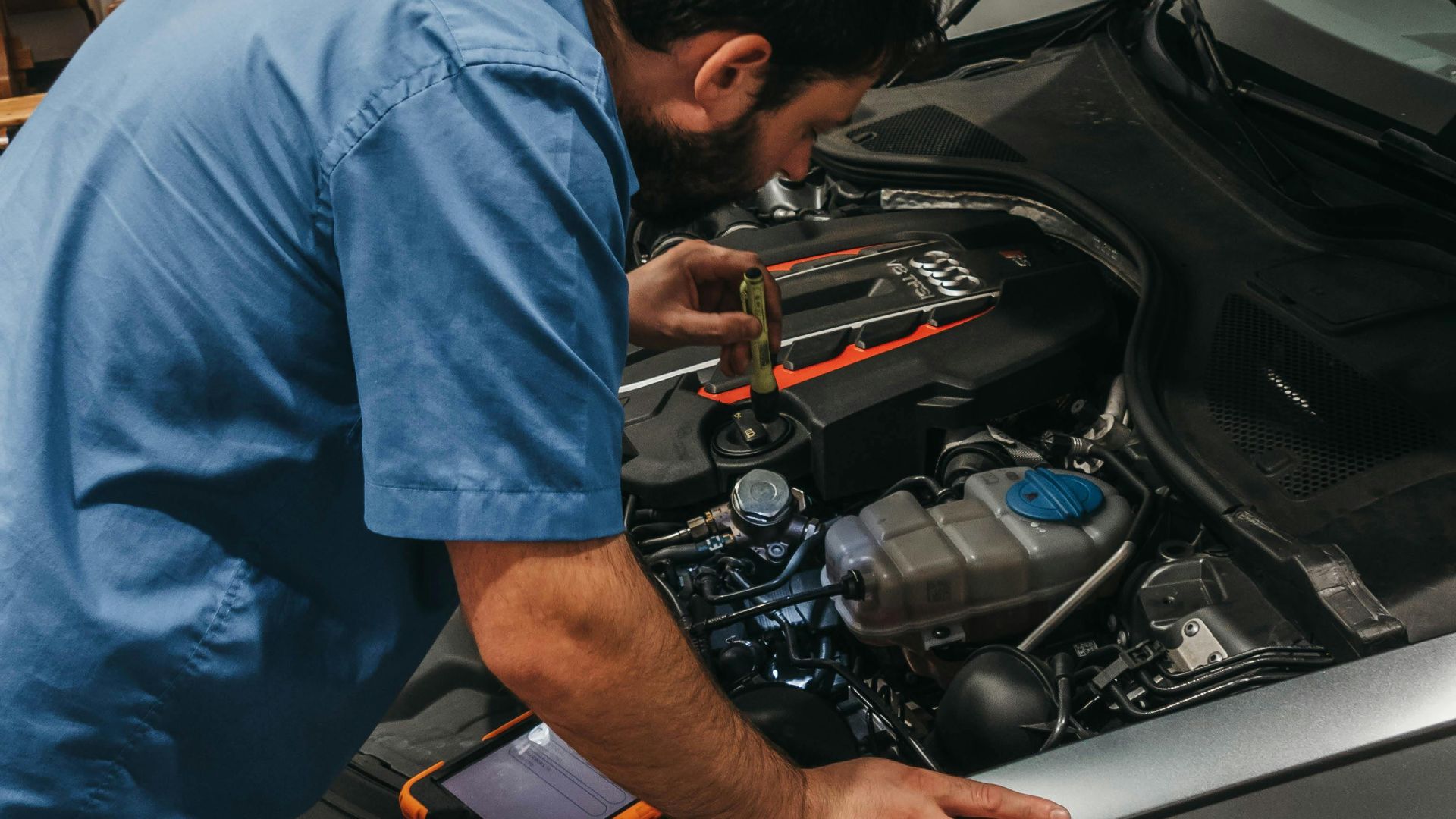 Mechanic in a workshop examining a car's engine under an open hood.
