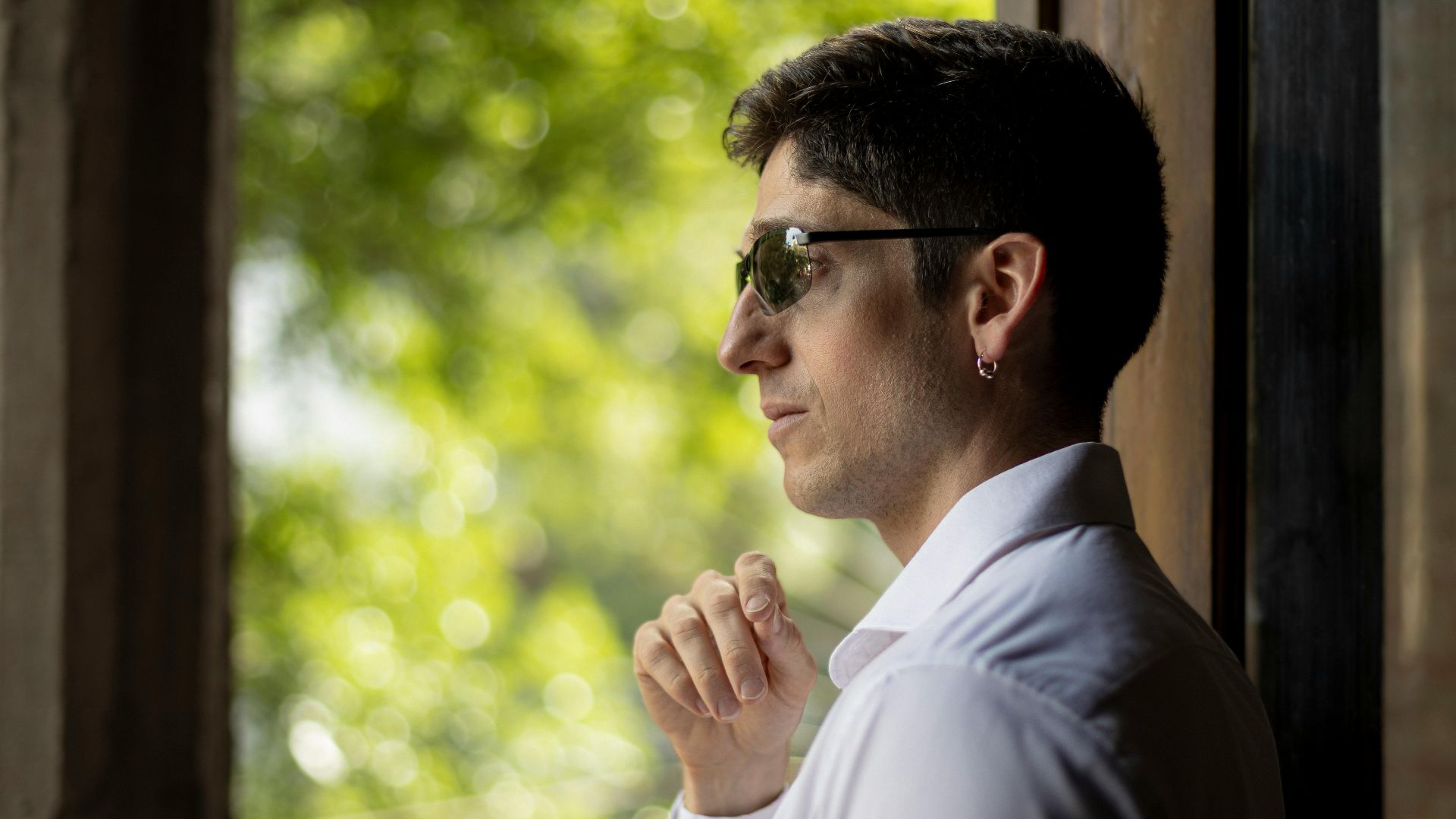 A stylish man wearing sunglasses gazes thoughtfully out a window, framed by greenery.