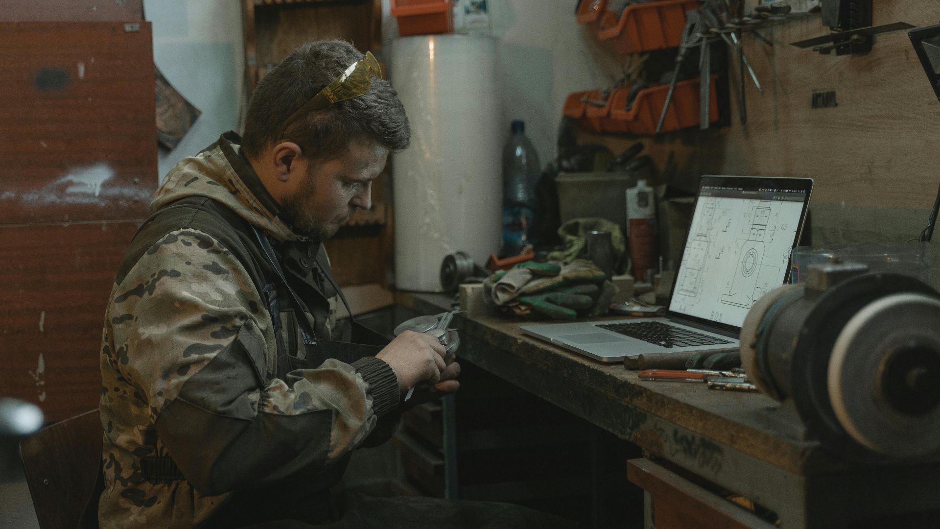 A man intensely focuses on repairing a part at his industrial workspace beside a laptop.