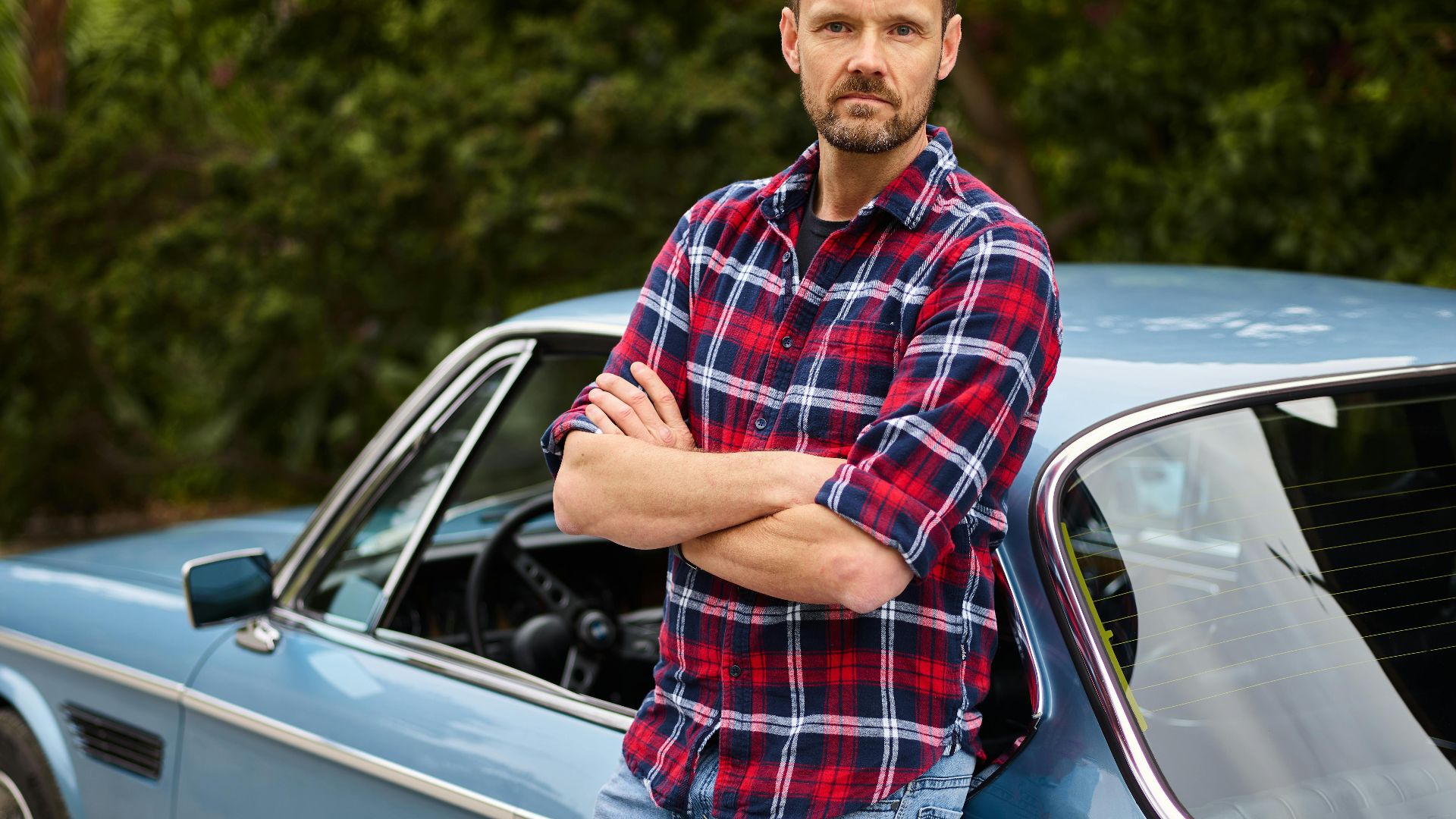 Adult bearded male in jeans and shirt standing with crossed arms leaned on blue retro auto near forest in daylight and looking at camera